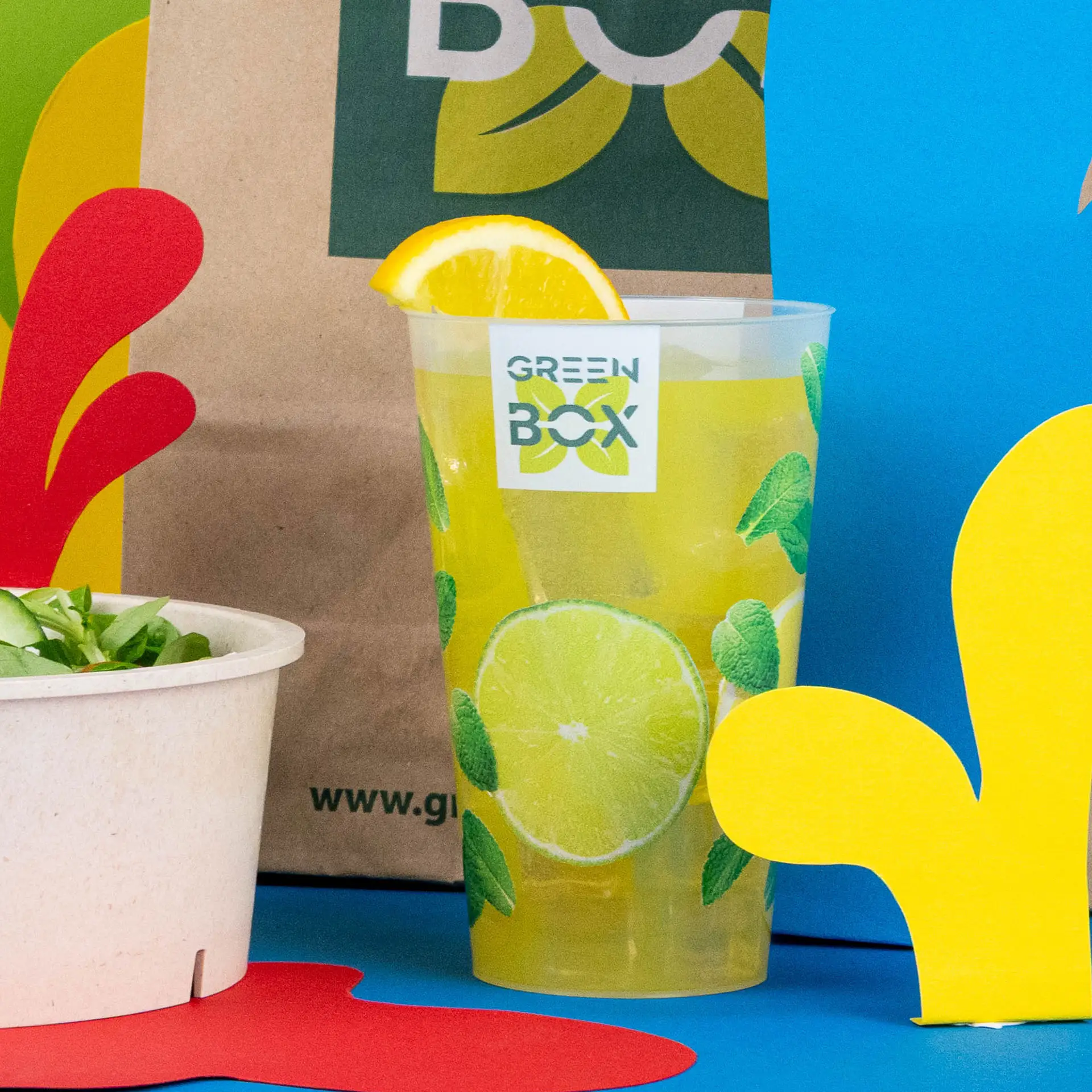Reusable cup with logo, 400 ml; golden-yellow beverage inside; accompanied by a burger and salad on a wooden table.