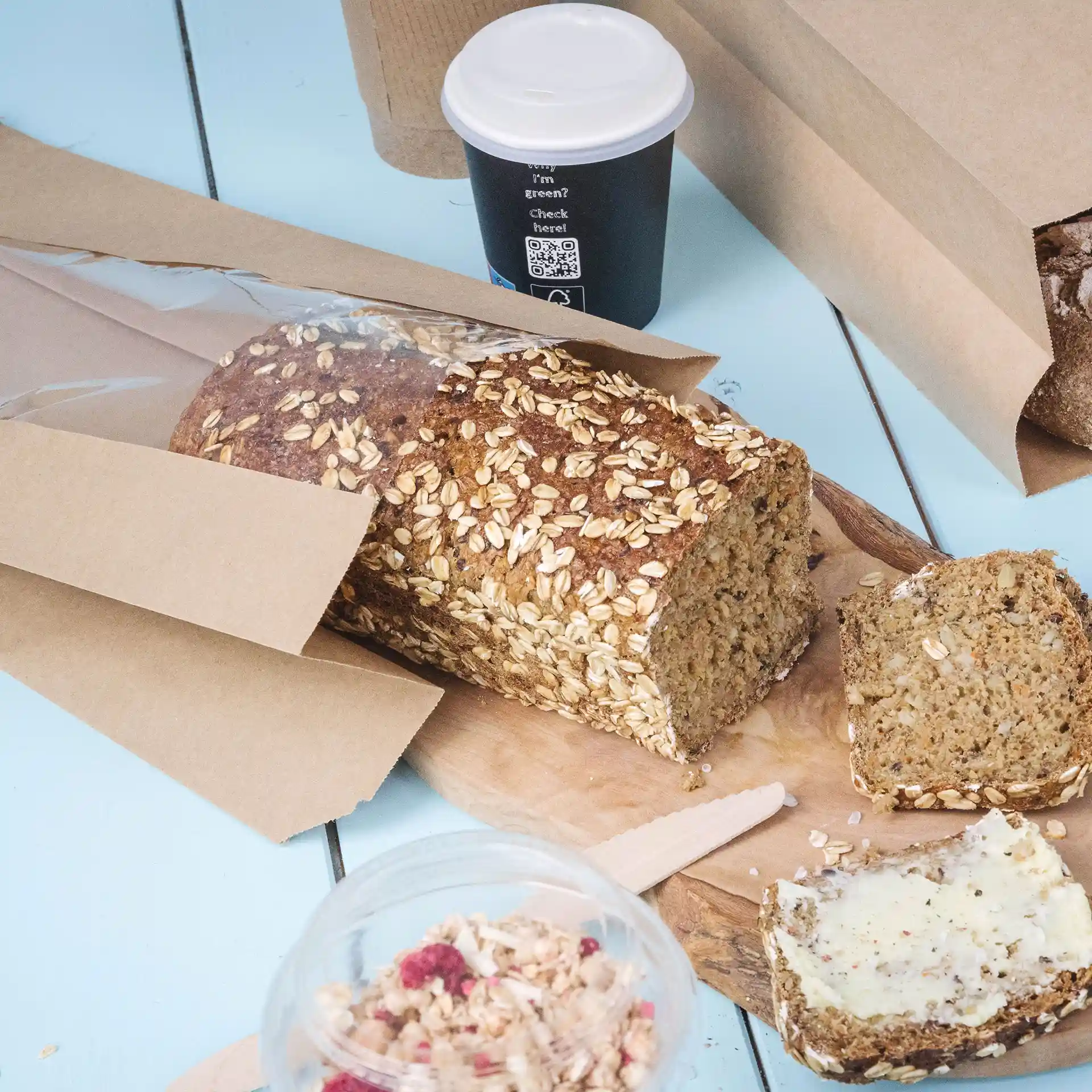 Bild eines Laibs Brot mit Haferflocken, teilweise geschnitten, auf einem Holzbrett mit einem Becher Kaffee und Granola in einem klaren Behälter.