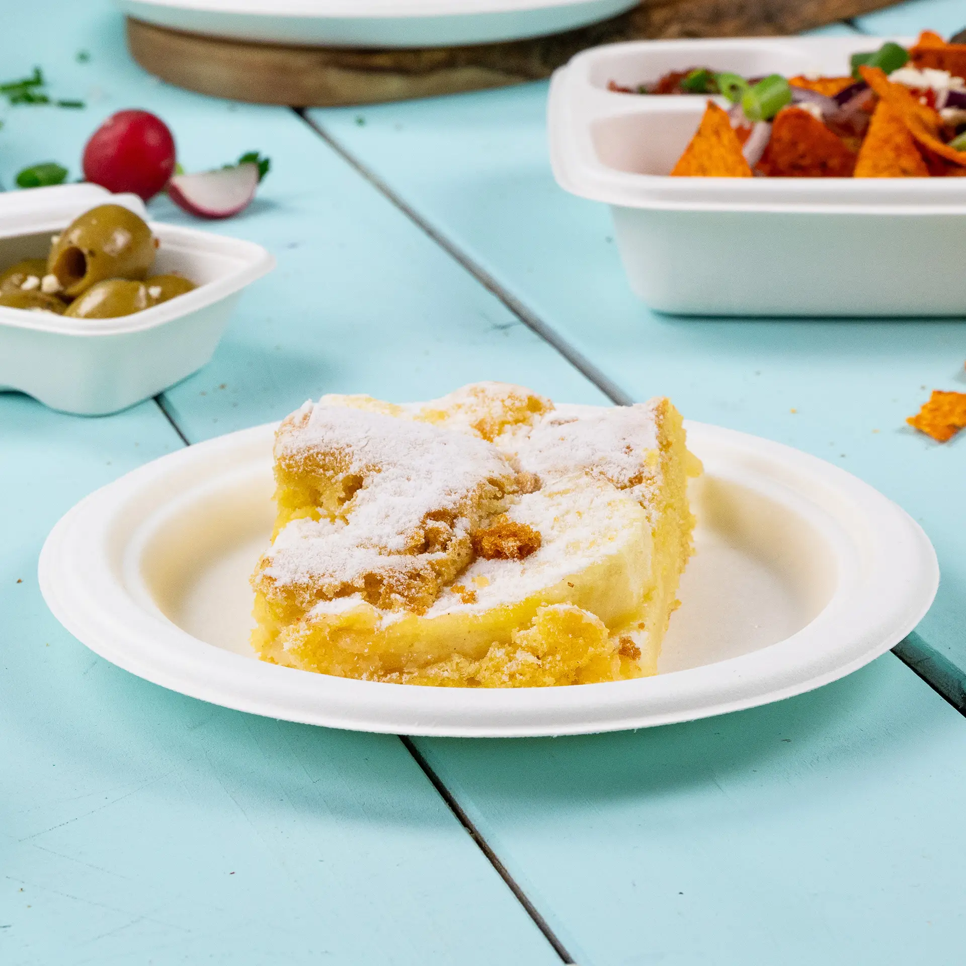A slice of cake dusted with powdered sugar on a round biodegradable sugarcane plate against a blue backdrop.