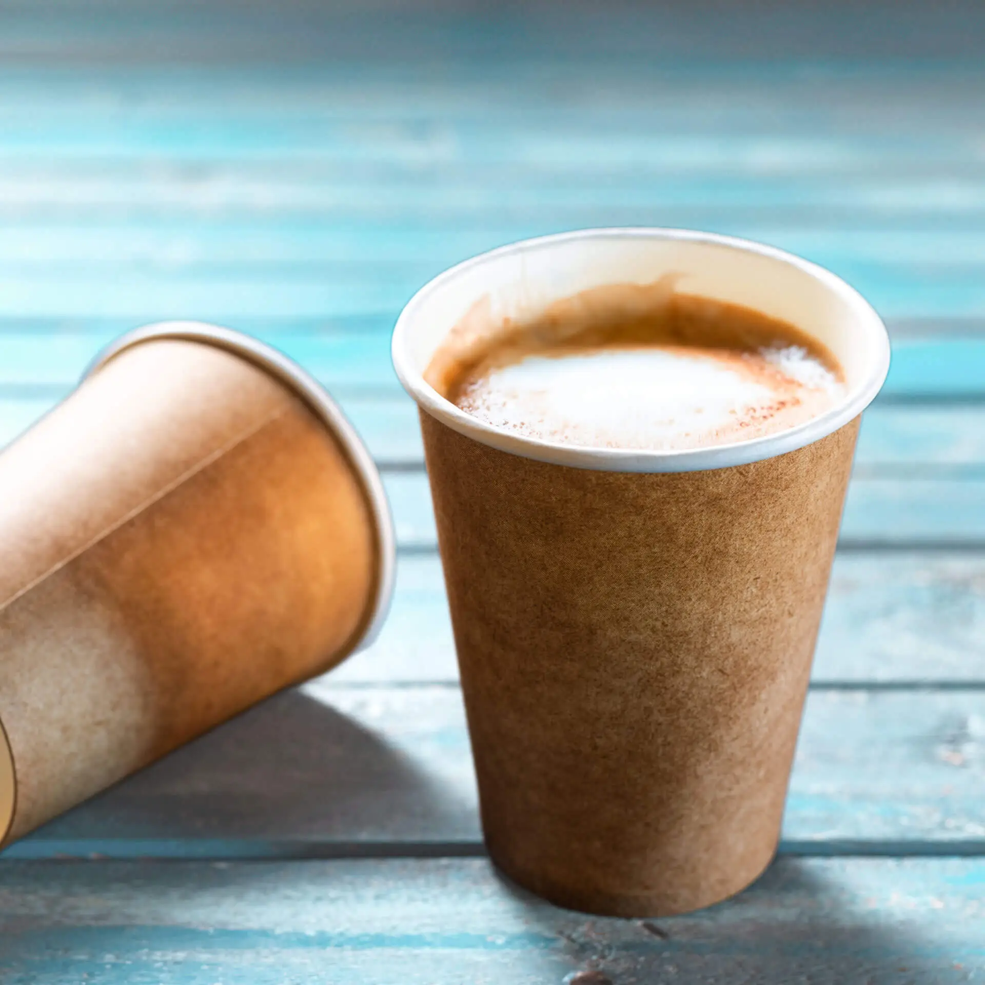 Single-wall brown paper cup with white inner lining, filled with coffee; another cup is featured behind it.