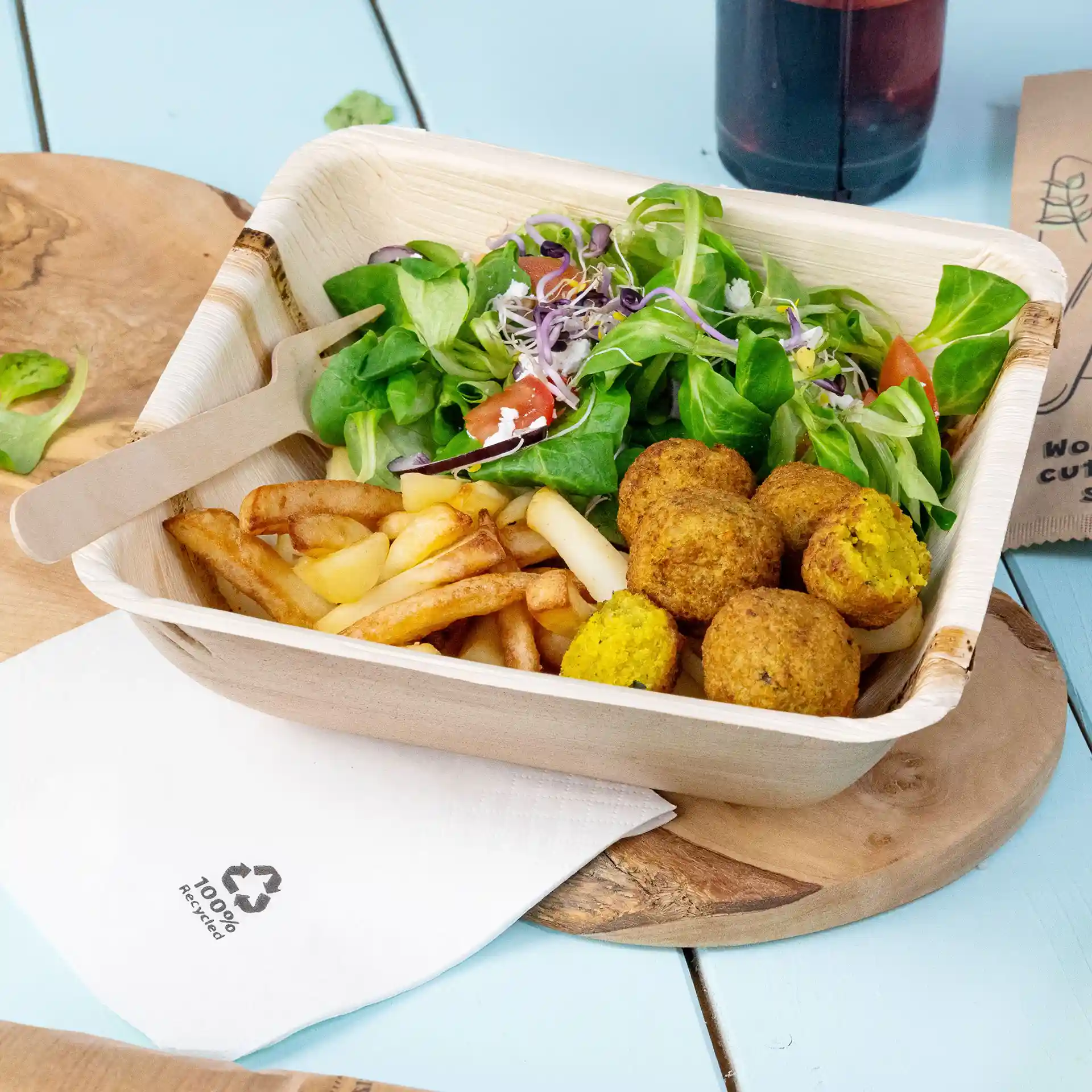 Tiefe quadratische Schale aus Palmblatt mit Salat, Pommes und Falafelbällchen, daneben eine Holzgabel und eine Serviette.