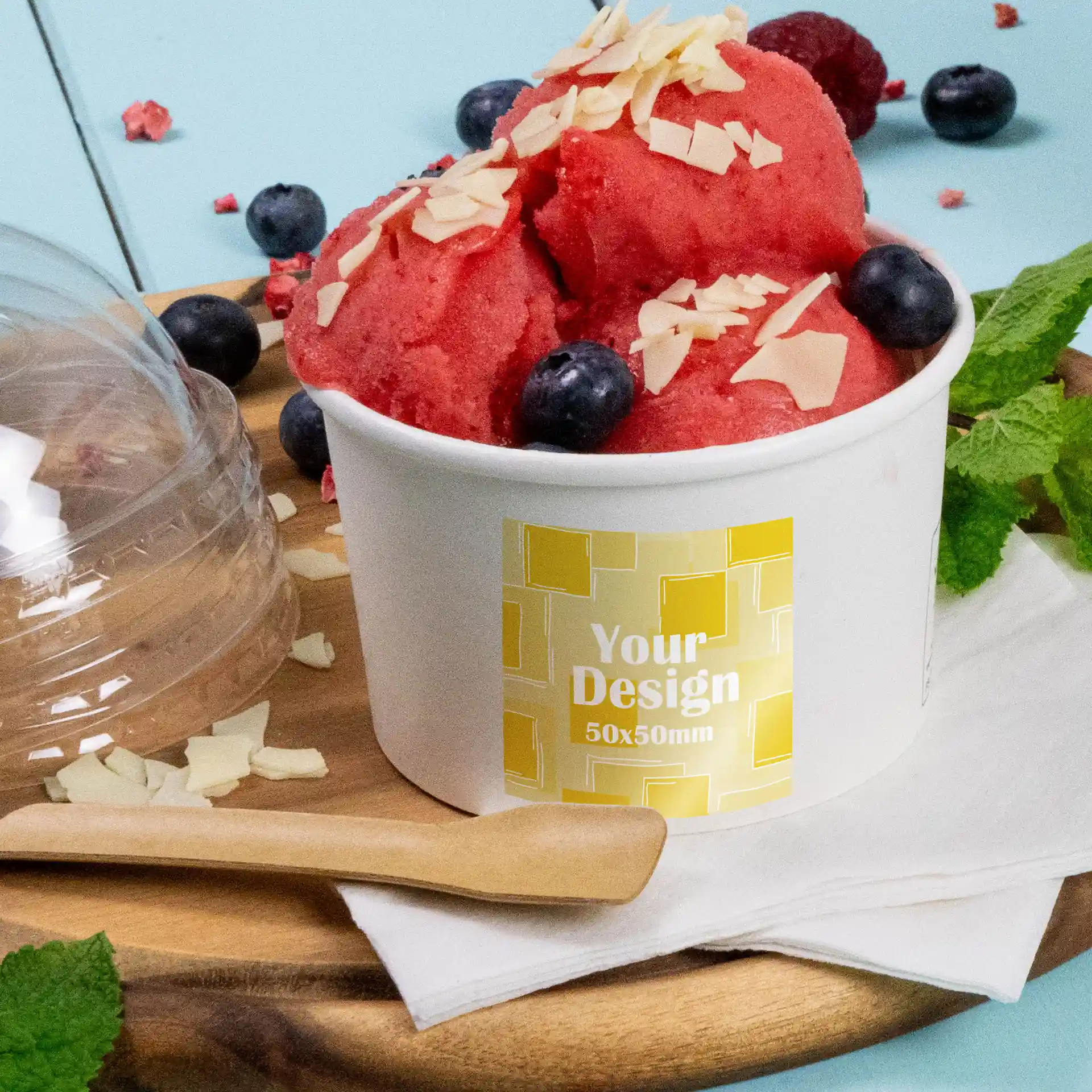 Cup of red sorbet topped with almond flakes and blueberries, with a printed label on a white container, placed on a wooden board.