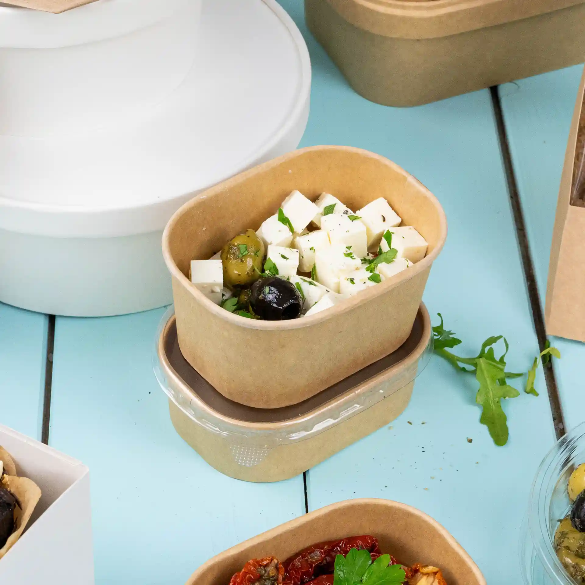 Rectangular brown cardboard food container with cheese and olives, placed on a blue surface with other containers in the background.
