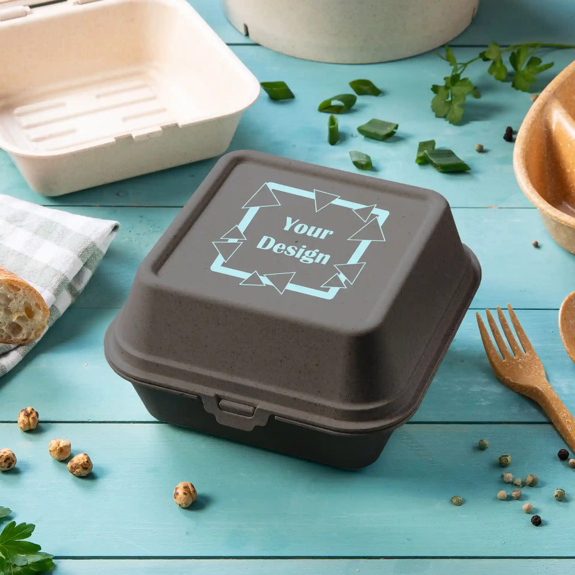Graue rechteckige Burgerbox mit Deckel, mit einem Designbereich, auf einer blauen Oberfläche mit Lebensmitteln drumherum.
