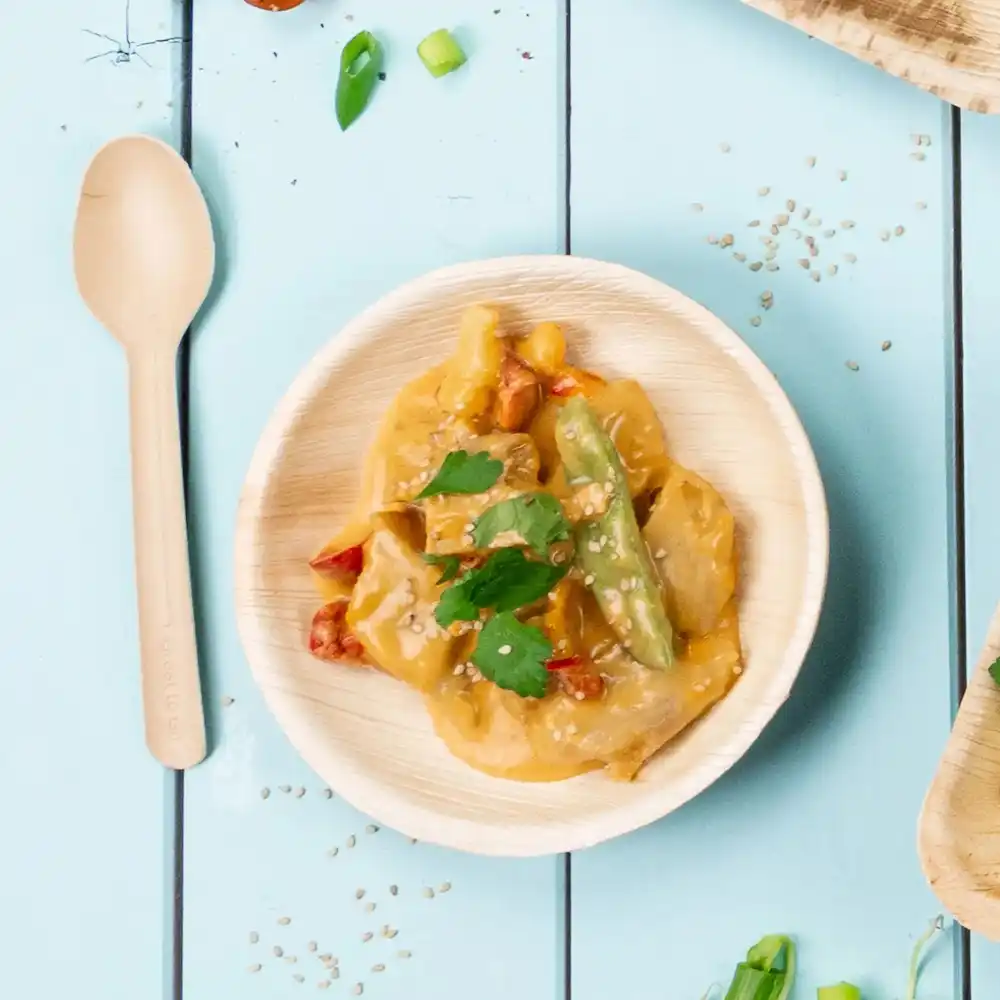 Bambusschüssel mit gebratenem Gemüse und Sauce, garniert mit Sesam und Koriander, neben einem Holzlöffel.