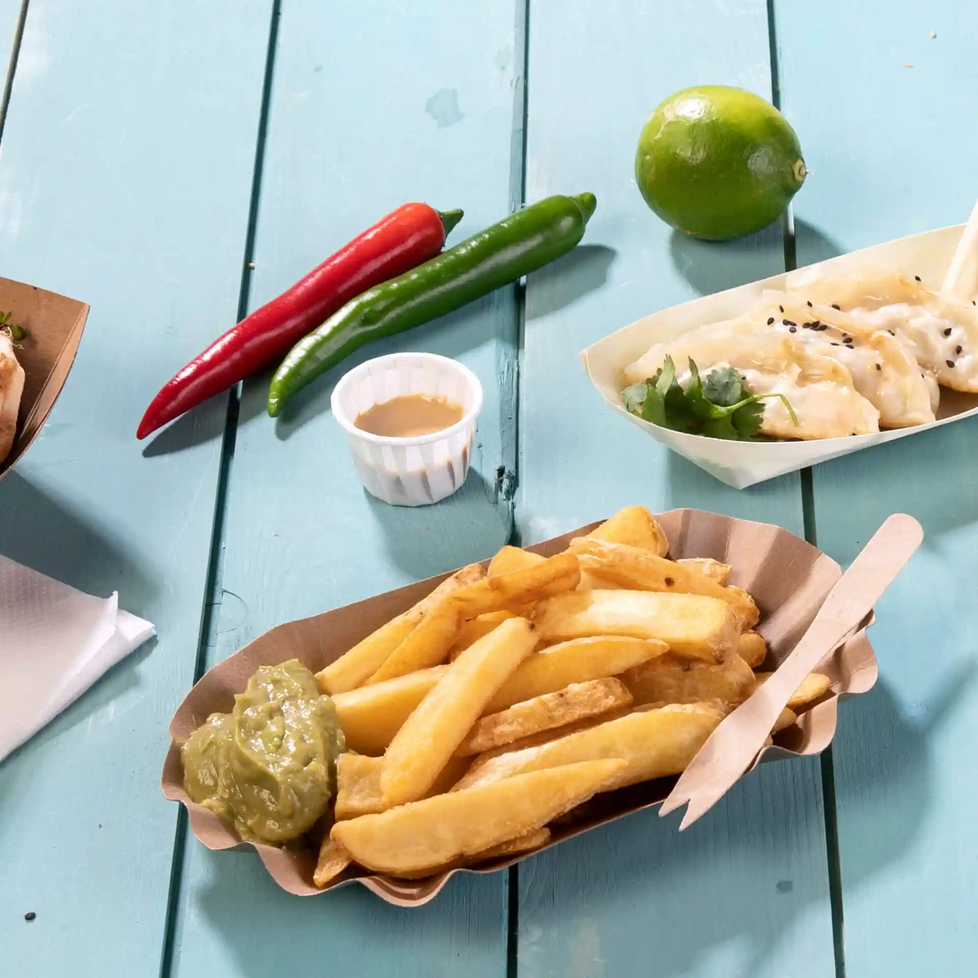 Bild einer Portion Pommes frites mit grüner Sauce in einer Papierschale, umgeben von Chilischoten, einer Limette und einem kleinen Becher Sauce.