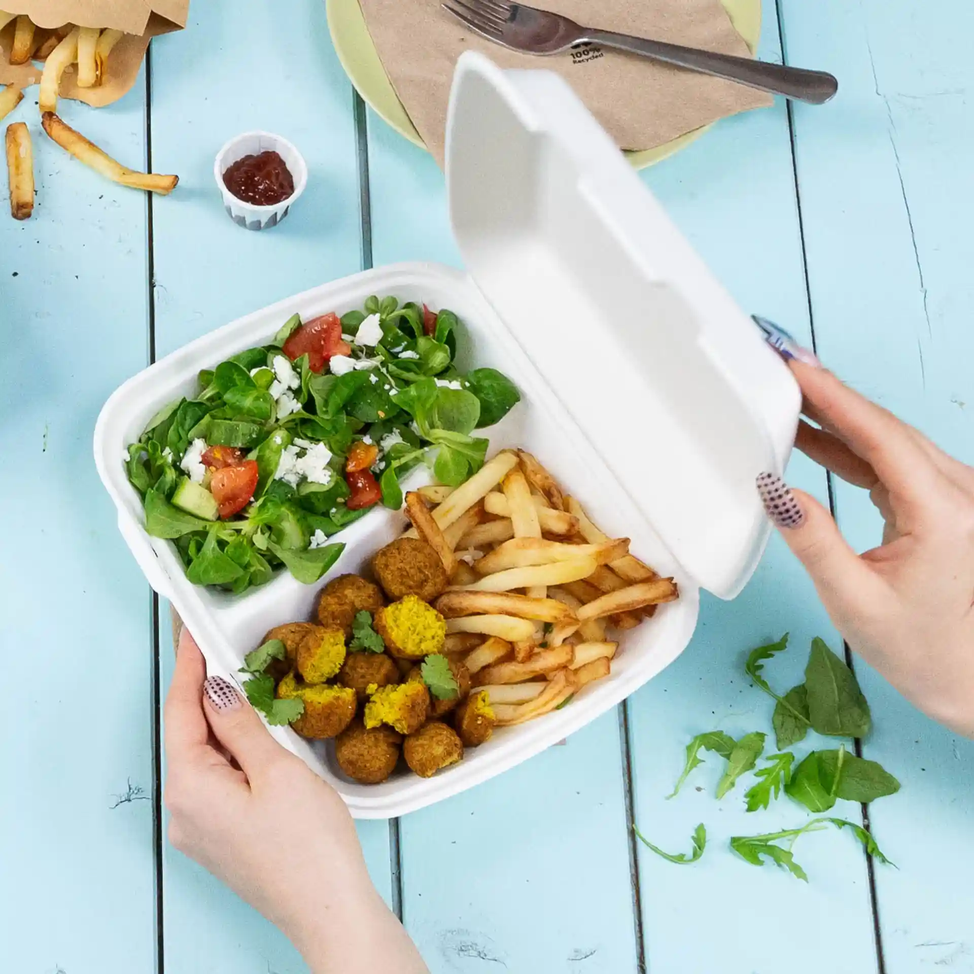 Eine Hand hält einen weißen Behälter mit zwei Fächern, gefüllt mit Salat, Pommes und Falafelbällchen.