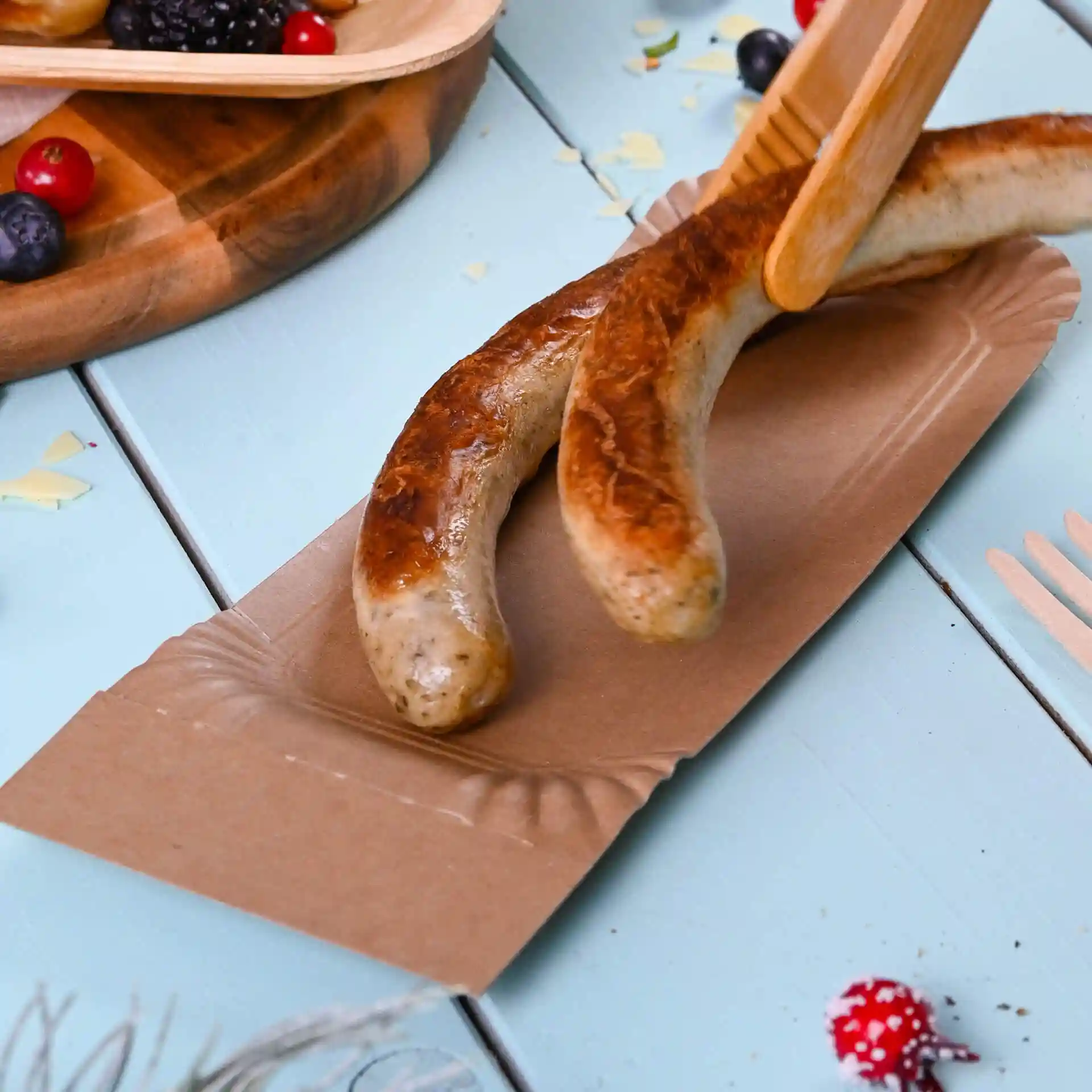 Zwei gegrillte Würstchen auf einem braunen Papptablett, auf einer hellblauen Fläche mit verstreuten Beeren.