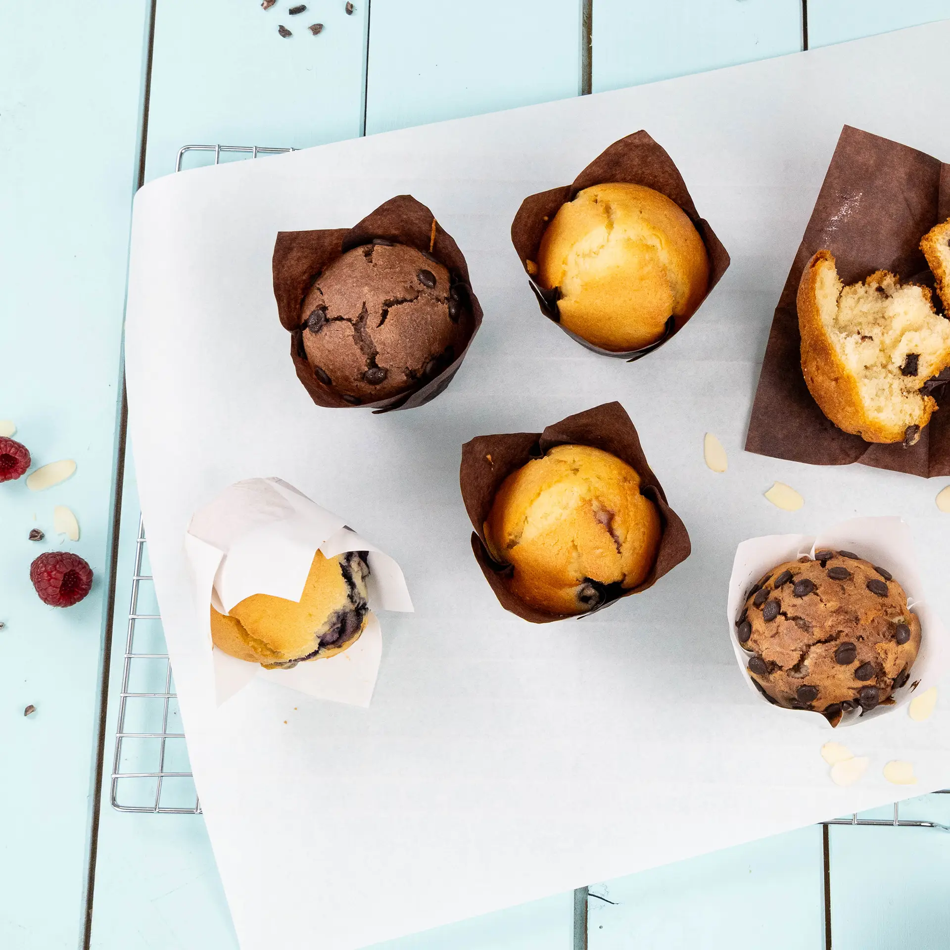 Assorted muffins in brown greaseproof paper cups on a white baking tray, with raspberries and almond flakes scattered nearby.