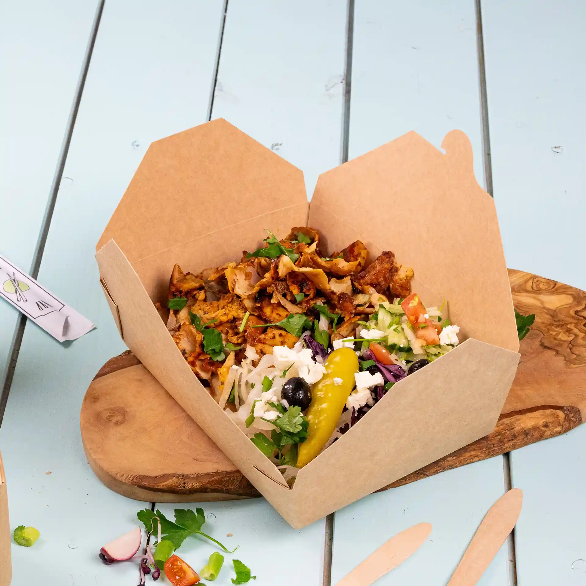 Brown cardboard takeaway container filled with food, including meat, salad, and a pickle, on a wooden board.