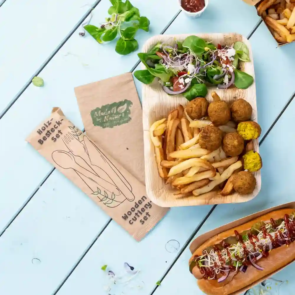 Holzbesteckset neben einem Essensplatten mit Salat, Pommes, Falafelbällchen und einem Hotdog auf einem blauen Tisch.