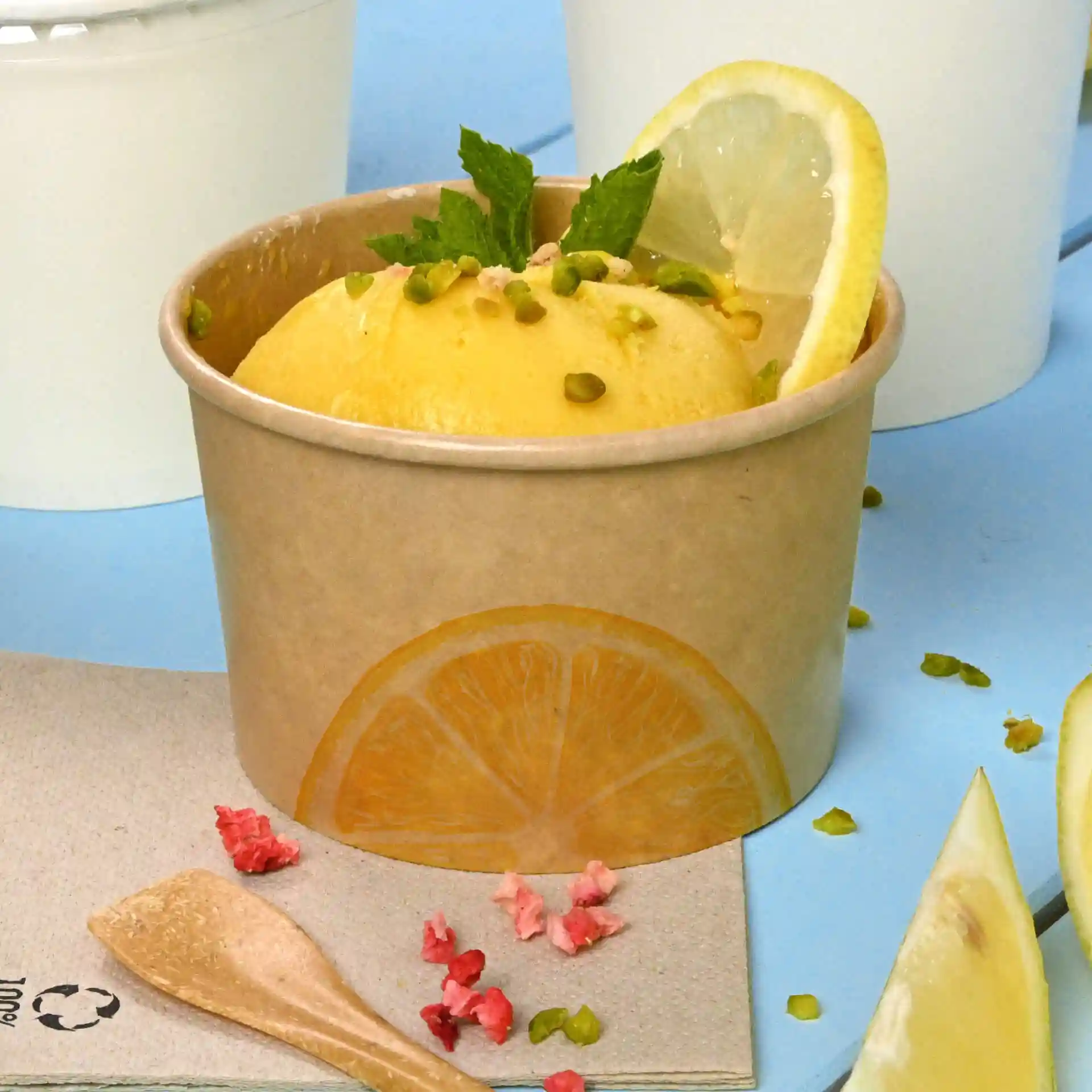 Brauner Pappbecher mit gelbem Eis, garniert mit Minzblättern und Zitronenscheiben, auf blauer Oberfläche.