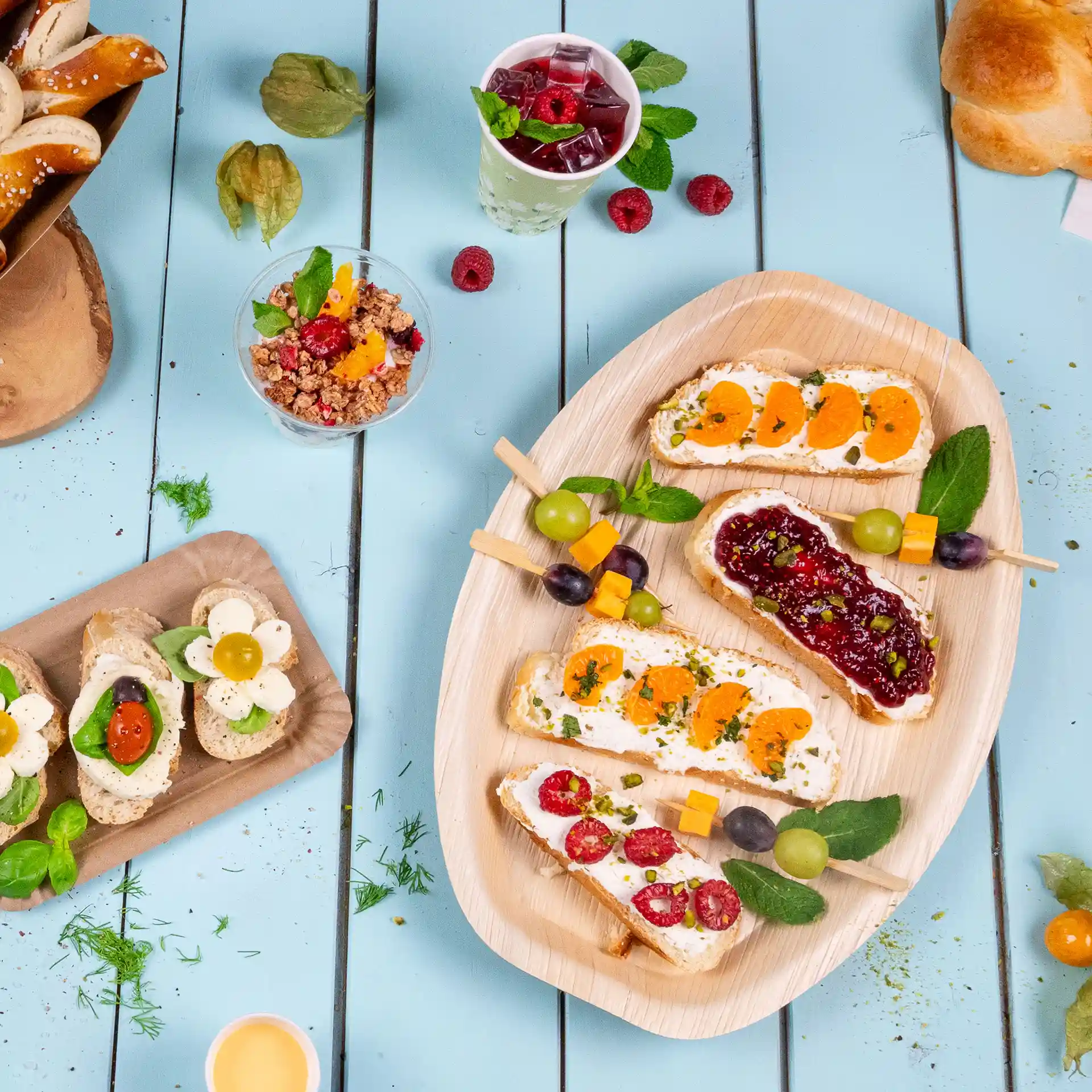 Rectangular palm leaf tray with assorted open-faced sandwiches, garnished with fruits and herbs, on a blue wooden table.