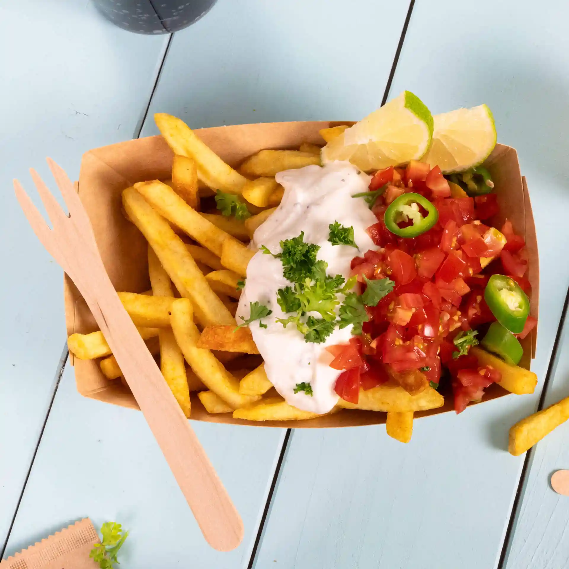 Wooden fork beside a serving of fries topped with sour cream, diced tomatoes, green peppers, and lime wedges.