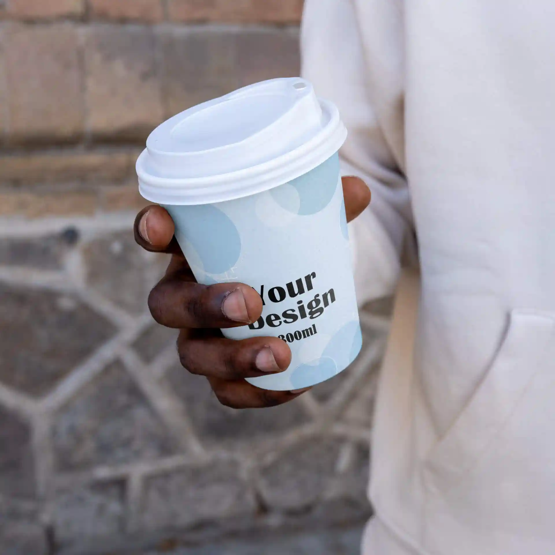 Eine Hand hält einen weißen Becher mit hellblauem Design und weißem Deckel, mit dem Text "Dein Design" und einem Volumen von 300 ml.
