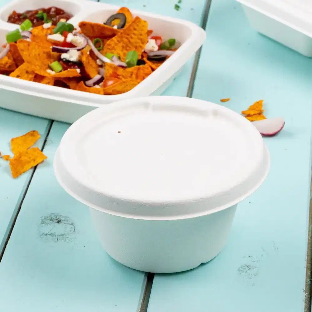 Weißer Papiersuppenbecher auf einem blauen Tisch mit einem Hintergrund aus Nachos und einem verschlossenen Lebensmittelbehälter.