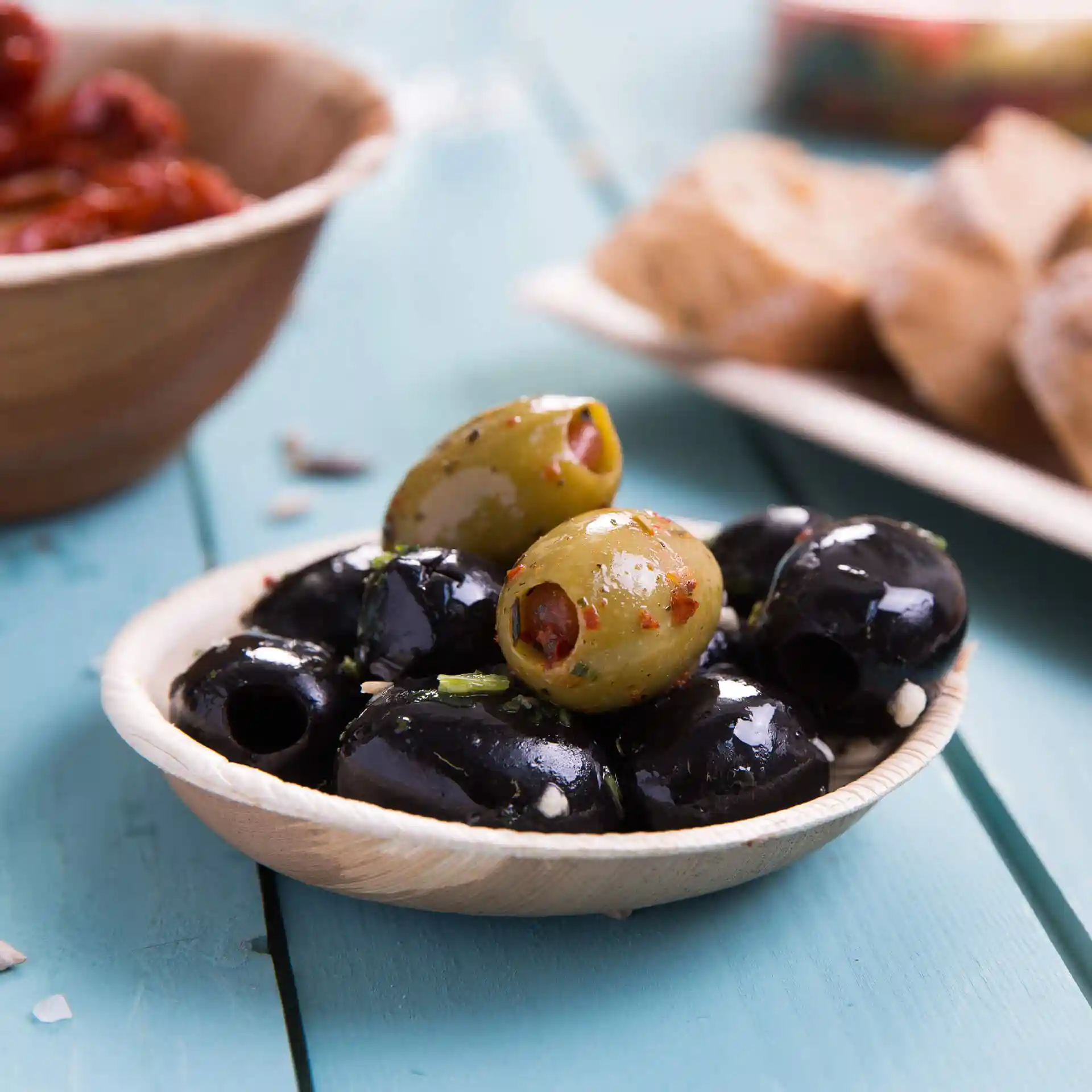 Bowl made of palm leaf containing black and green olives, placed on a blue wooden surface.
