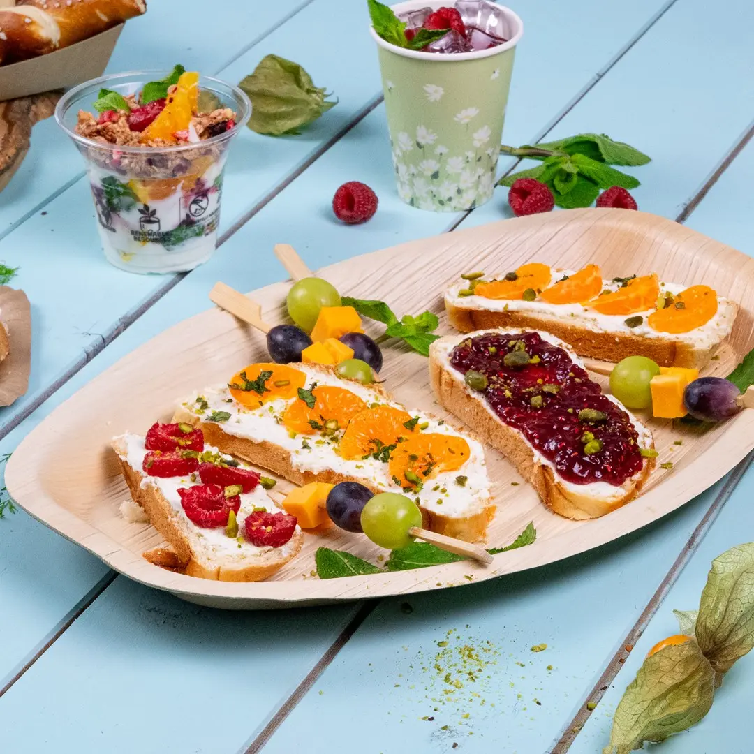 Platte mit verschiedenen Fingerfood, darunter Brot mit Frischkäse, Obst und einem Obstspieß, sowie einem Joghurt-Parfait.