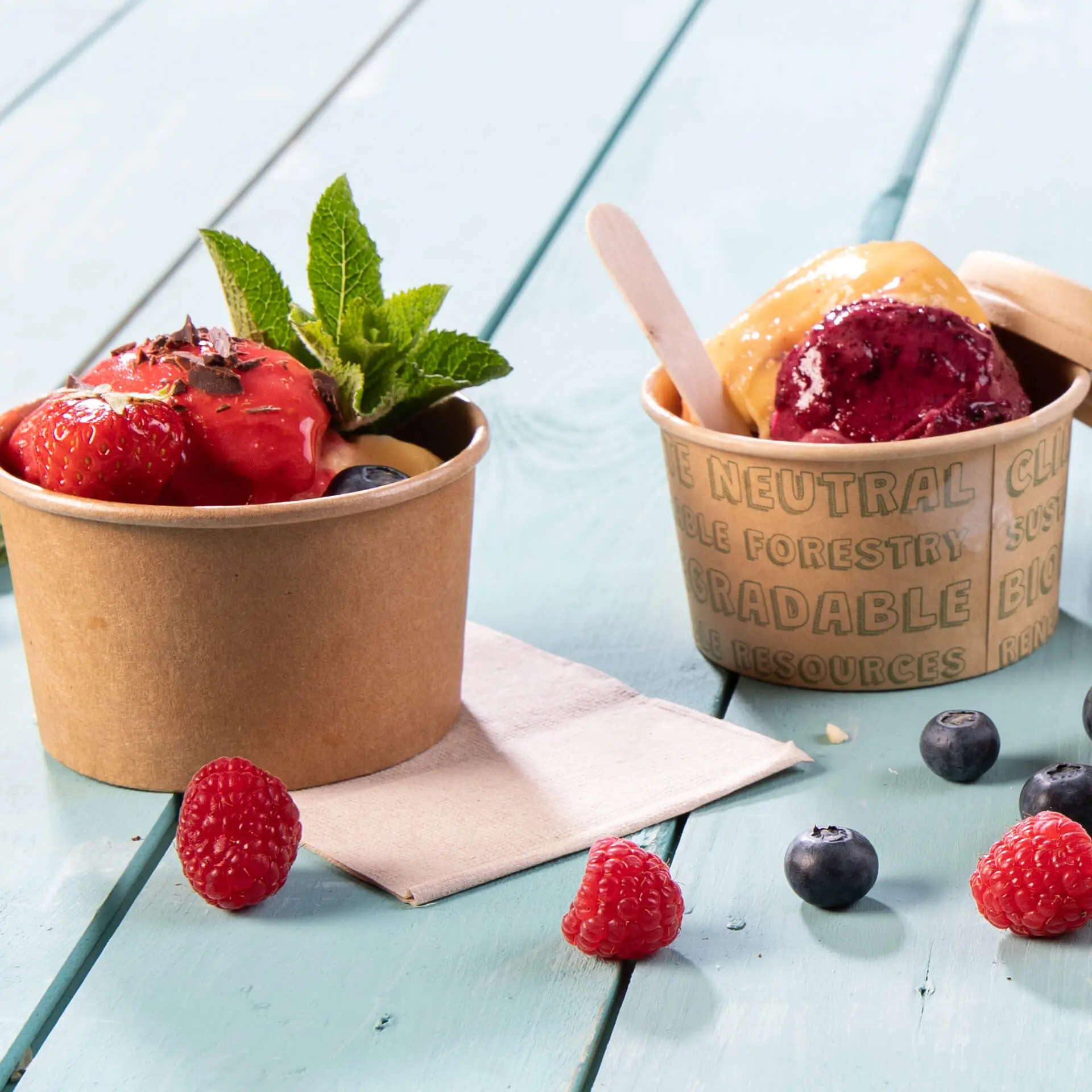Two paper cups with ice cream and fruit toppings, one with strawberries and mint, the other with purple and yellow ice cream.