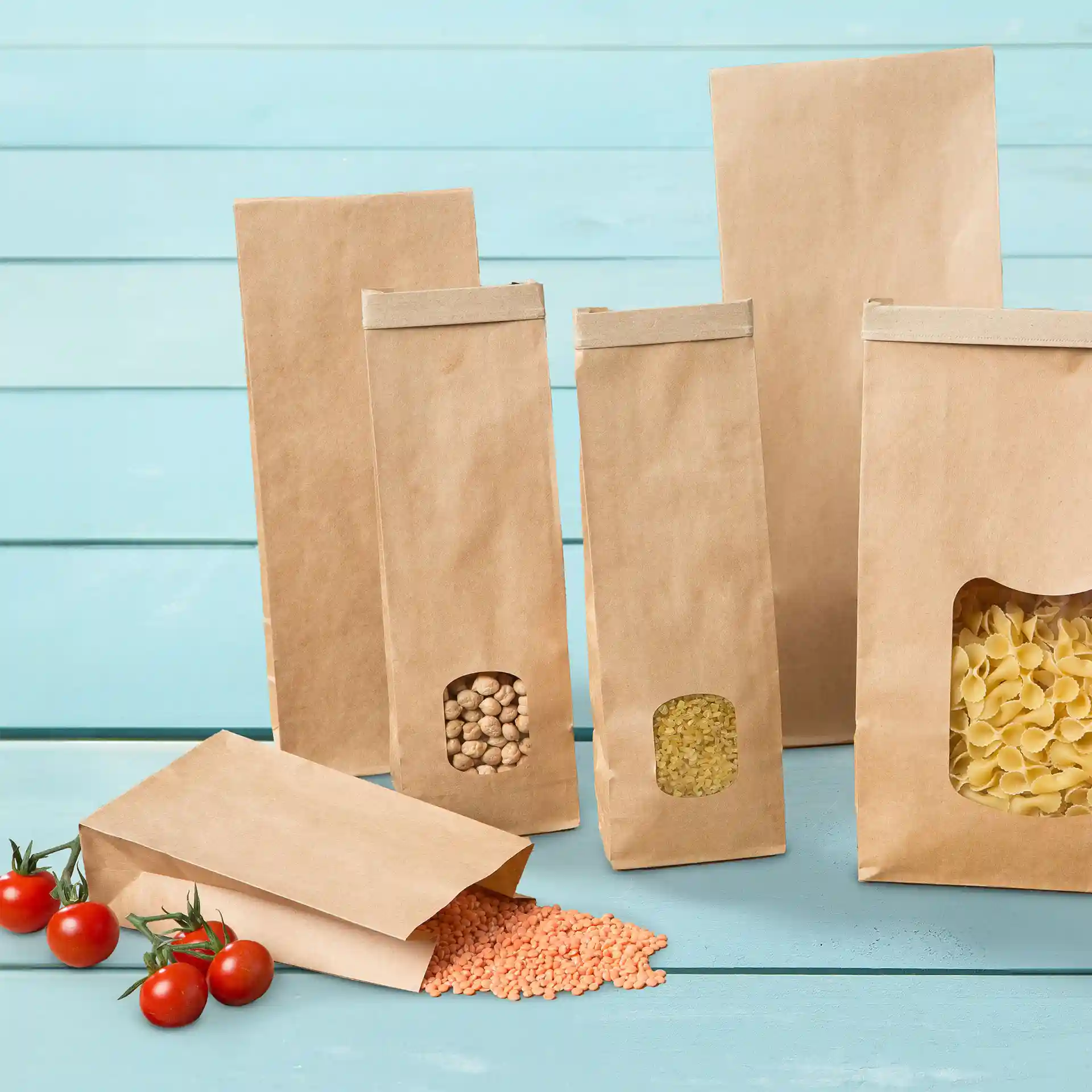 Braune Kraftpapiertüten in verschiedenen Größen mit schlichtem Design, einige enthalten Linsen und Pasta, auf blauem Hintergrund.