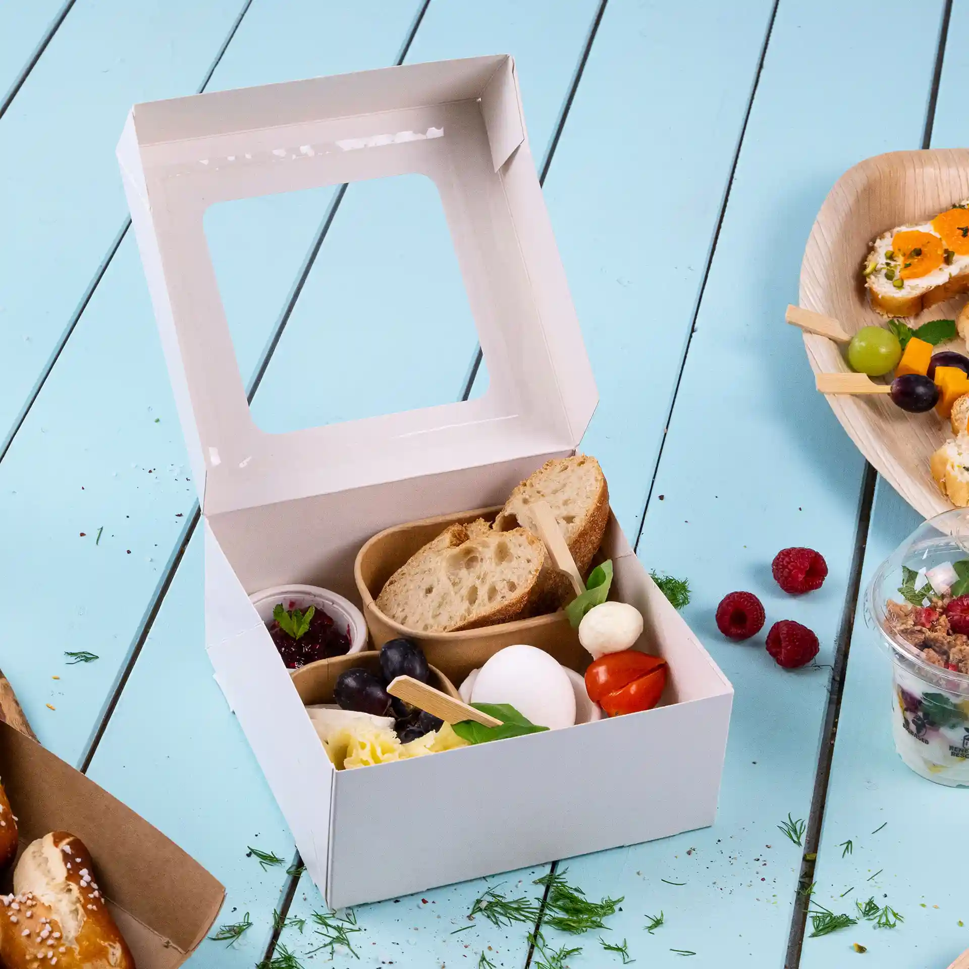 Weißer Karton mit Zellulosefenster, gefüllt mit Brot, Käse und verschiedenen Früchten.