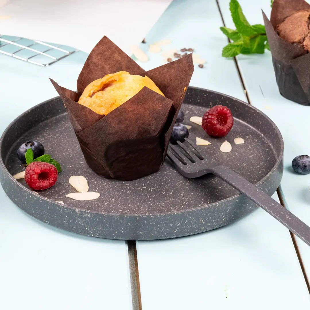 Muffin in a brown paper wrapper on a gray plate with raspberries, blueberries, almond slices, and a small fork.