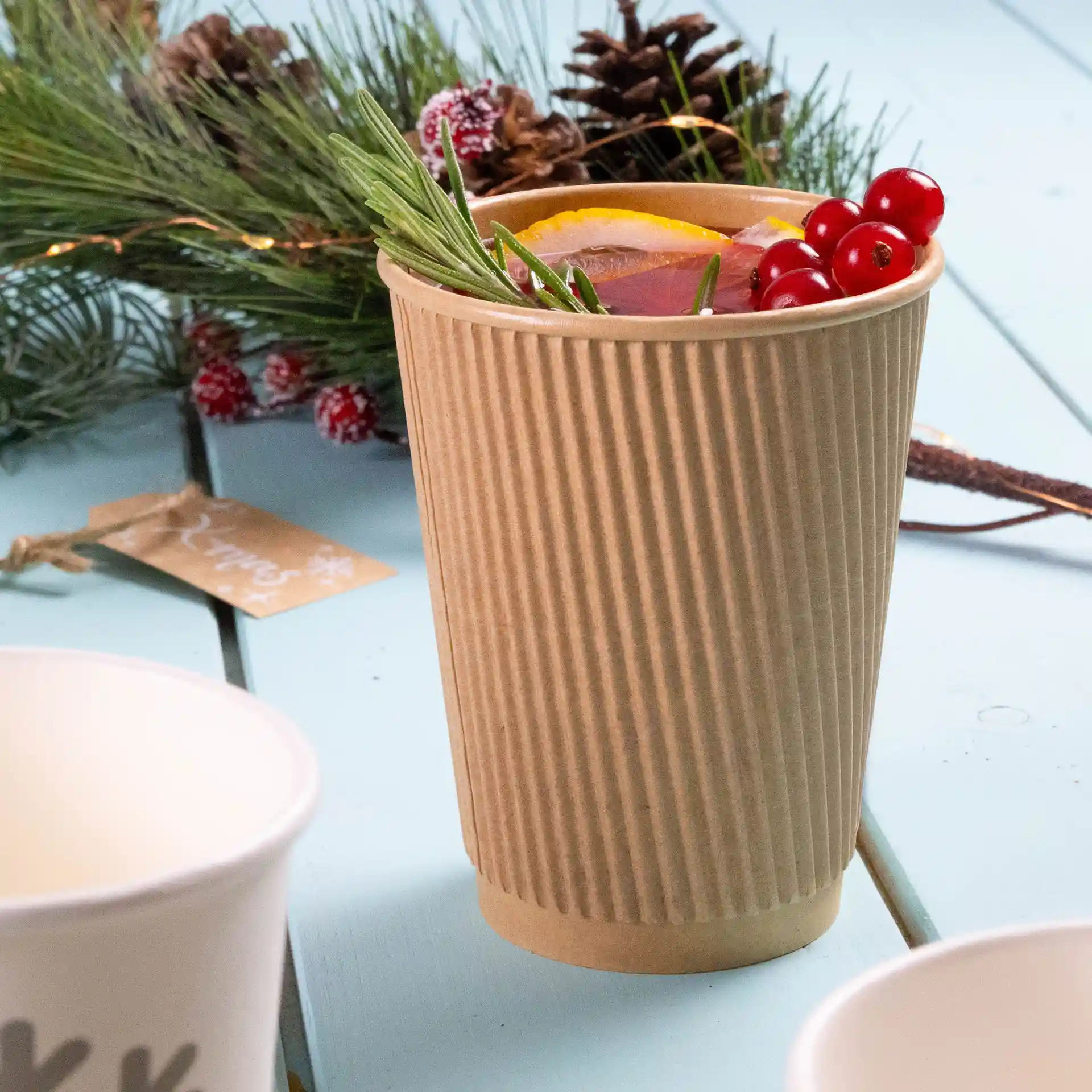 Brown ribbed paper cup filled with a drink, garnished with orange, berries, and herbs, on a light blue surface.