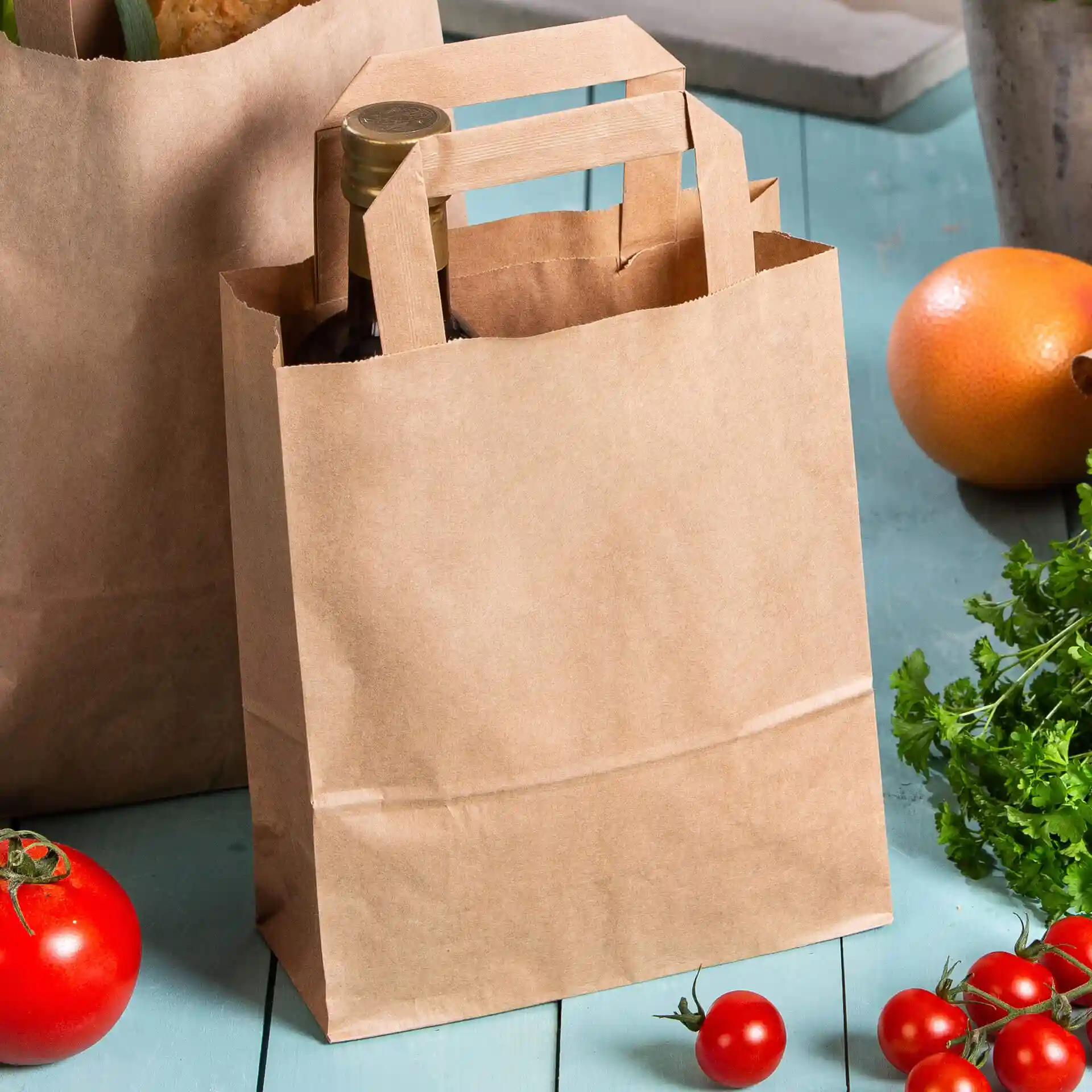 Brown paper carrier bag with handles, containing a bottle, surrounded by tomatoes and other produce.