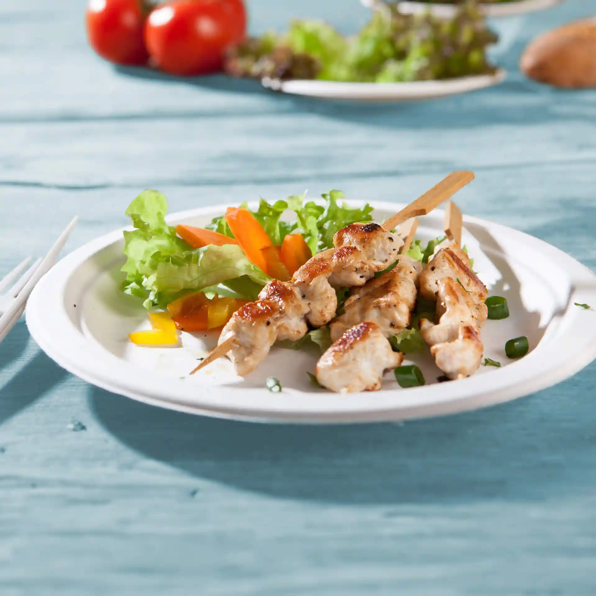 Runde Zuckerröhrenplatte mit gegrillten Hähnchenspießen und gemischtem Salat, auf einer blauen Holzoberfläche.