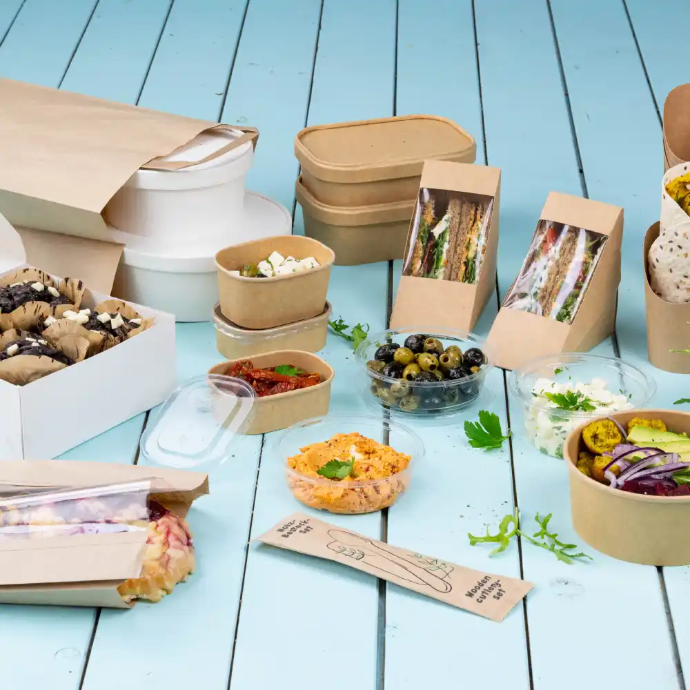 Eine Auswahl an Lebensmittelbehältern umfasst Papierboxen, Schalen und durchsichtige Plastikbecher mit verschiedenen Gerichten und Snacks.