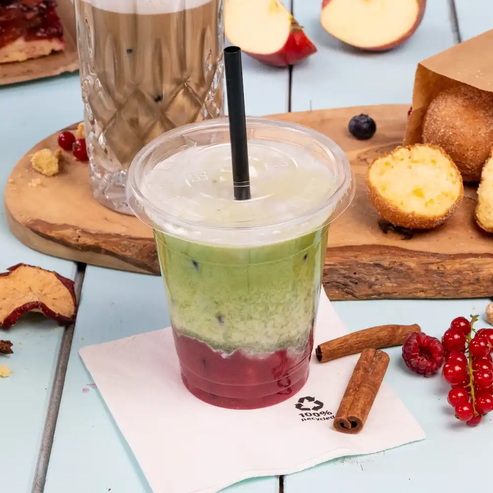 Eisgekühltes Schichtgetränk in einem transparenten Becher mit einem Strohhalm, umgeben von Snacks und einer Serviette auf einer hellblauen Oberfläche.