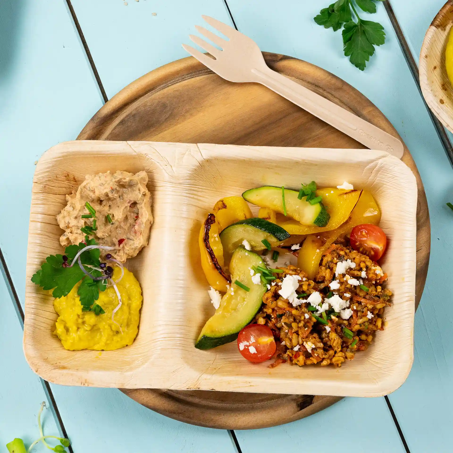 Rechteckiger Teller aus Palmblatt mit zwei Fächern, gefüllt mit Gemüse, Getreide und Dips, auf einer Holzoberfläche.