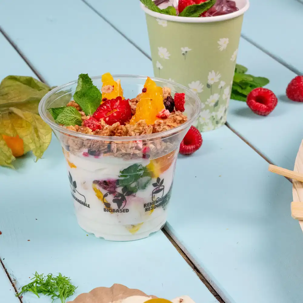 Klarer Plastikdessertbecher gefüllt mit Joghurt, Früchten und Granola, auf einer blauen Oberfläche mit Minzblättern.