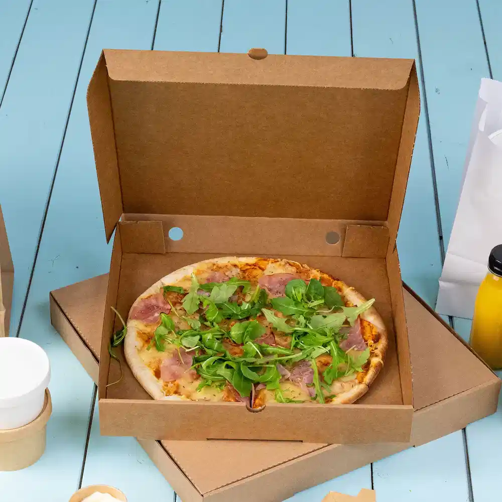Pizza in einer Kartonbox mit Schinken und gemischtem Grün auf einer blauen Holzoberfläche.
