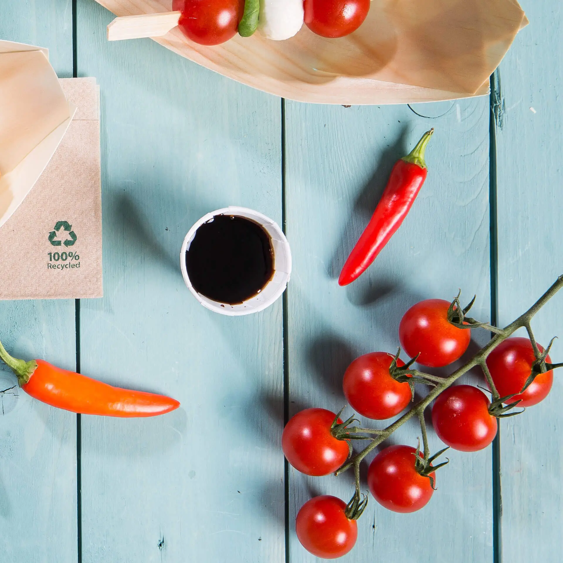 Bild eines Holzbretts mit Kirschtomaten, Chilischoten, einem kleinen weißen Papierbecher mit dunkler Sauce und einer braunen Verpackung mit Recycling-Symbol.