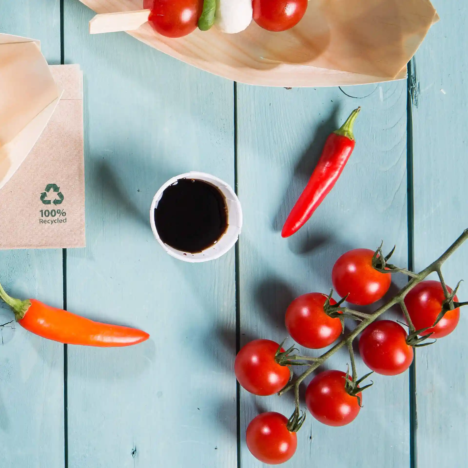 Image of a paper cup with dark liquid, surrounded by cherry tomatoes, chili peppers, and a wooden plate on a blue wooden surface.
