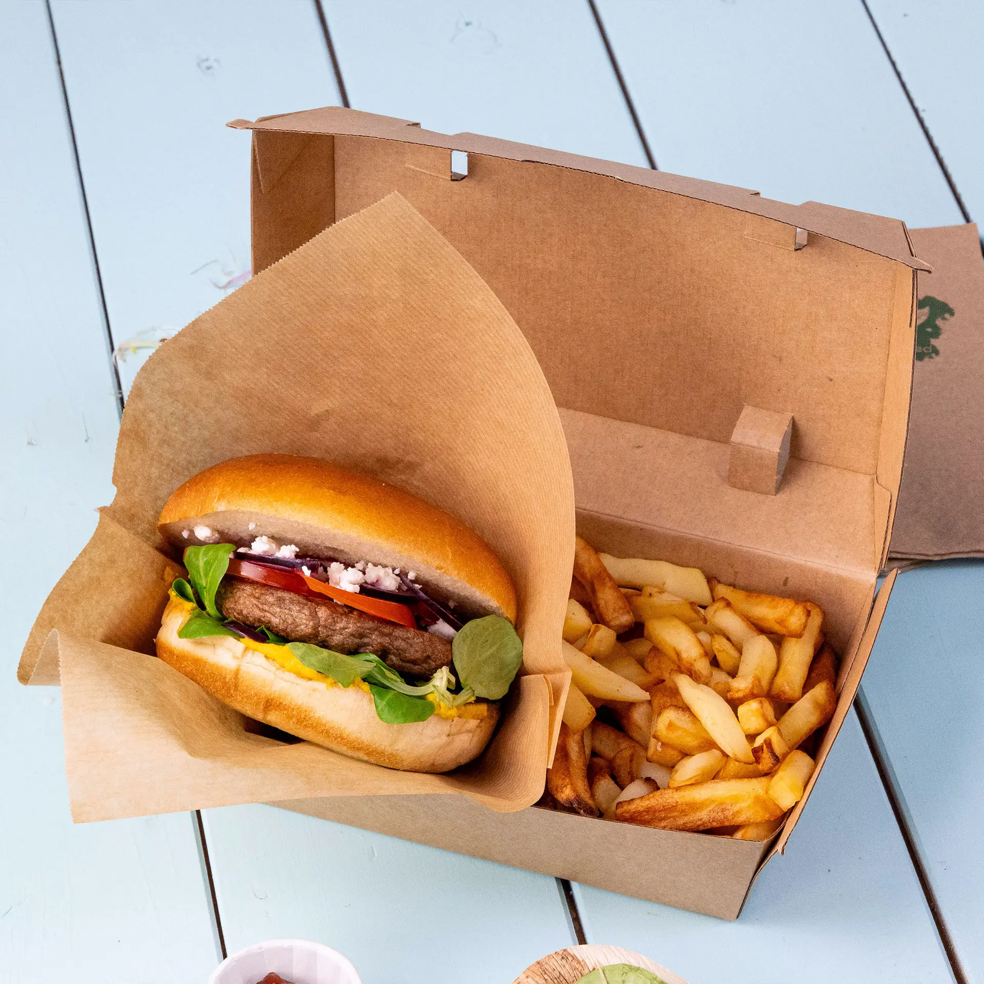 A takeaway box made of brown kraft cardboard with a burger and fries inside. The box has a high flip lid and is biodegradable.