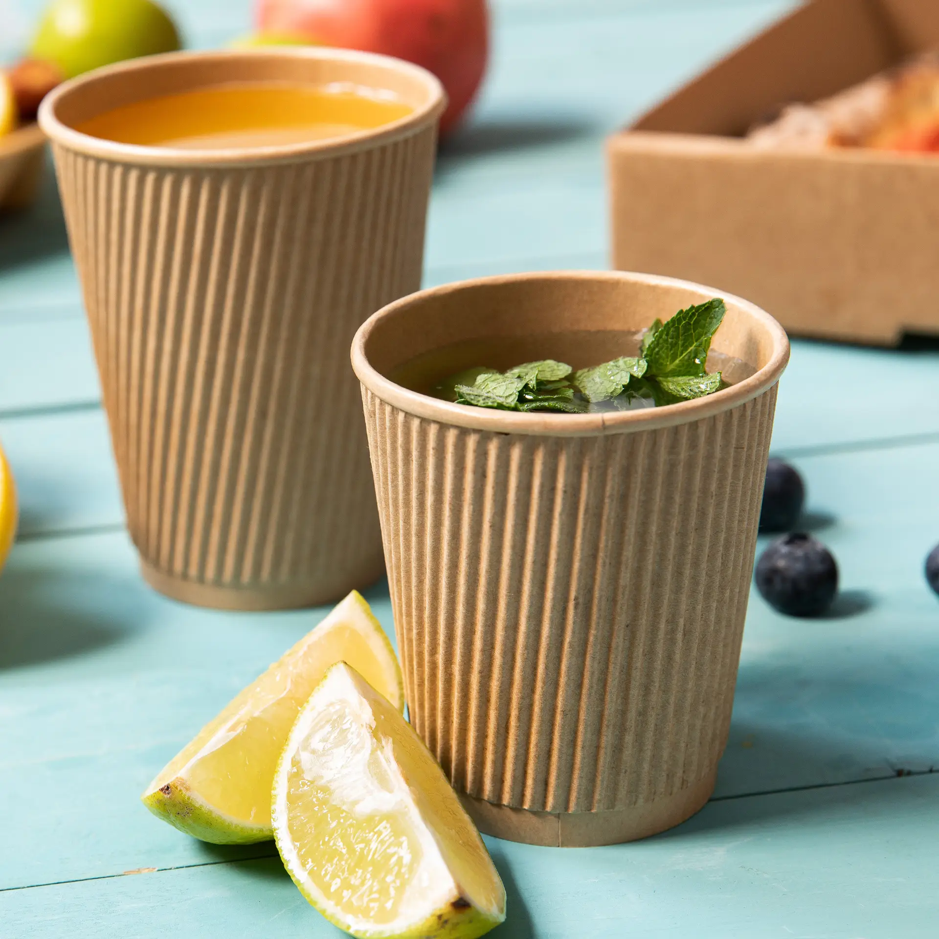 Two brown, ribbed, insulated disposable cups for hot drinks, one with mint leaves, and lime wedges beside them.