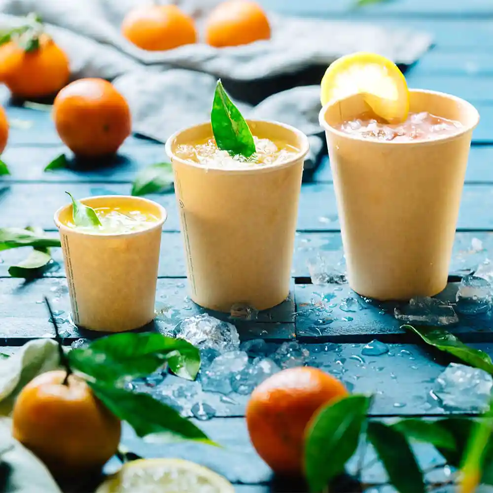 Three brown paper cups filled with beverages, placed on a blue wooden surface with ice and citrus fruits around.