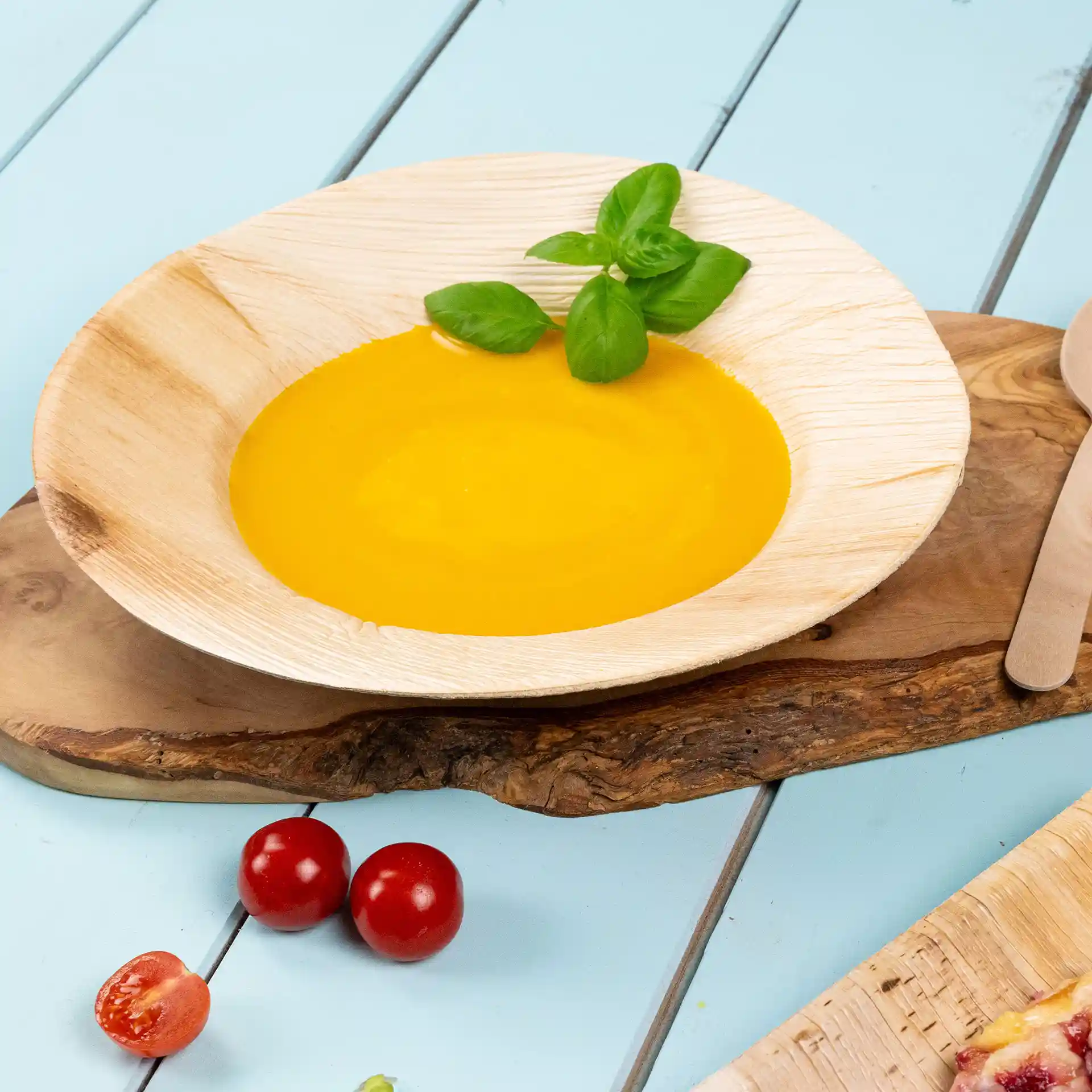 Tiefe runde Teller aus Palmblatt mit gelber Suppe und Basilikumblättern, auf einer Holzoberfläche mit Kirschtomaten daneben.