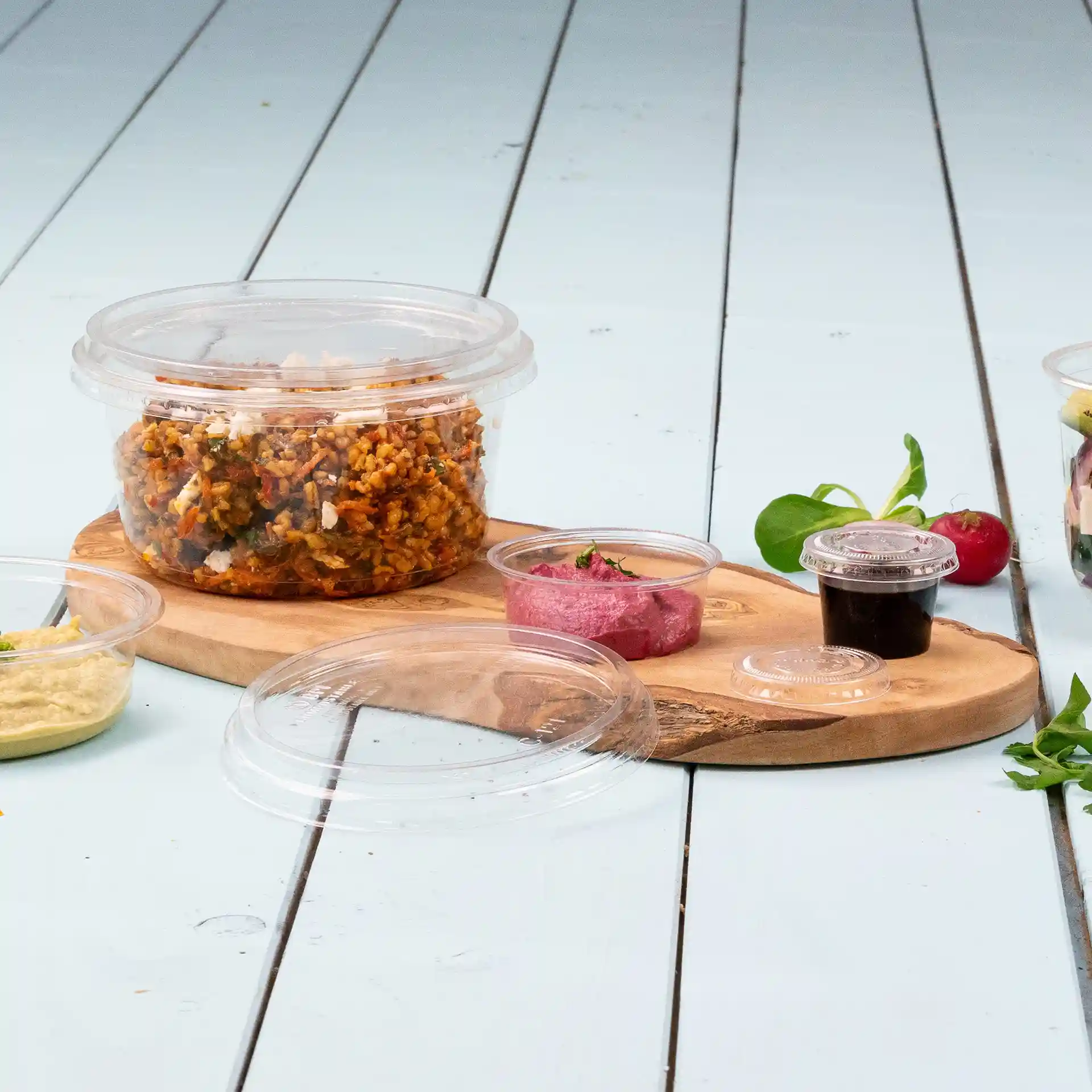 Transparenter runder Deli-Behälter mit Deckel, gefüllt mit Essen, auf einem Holzbrett mit kleinen Bechern daneben.