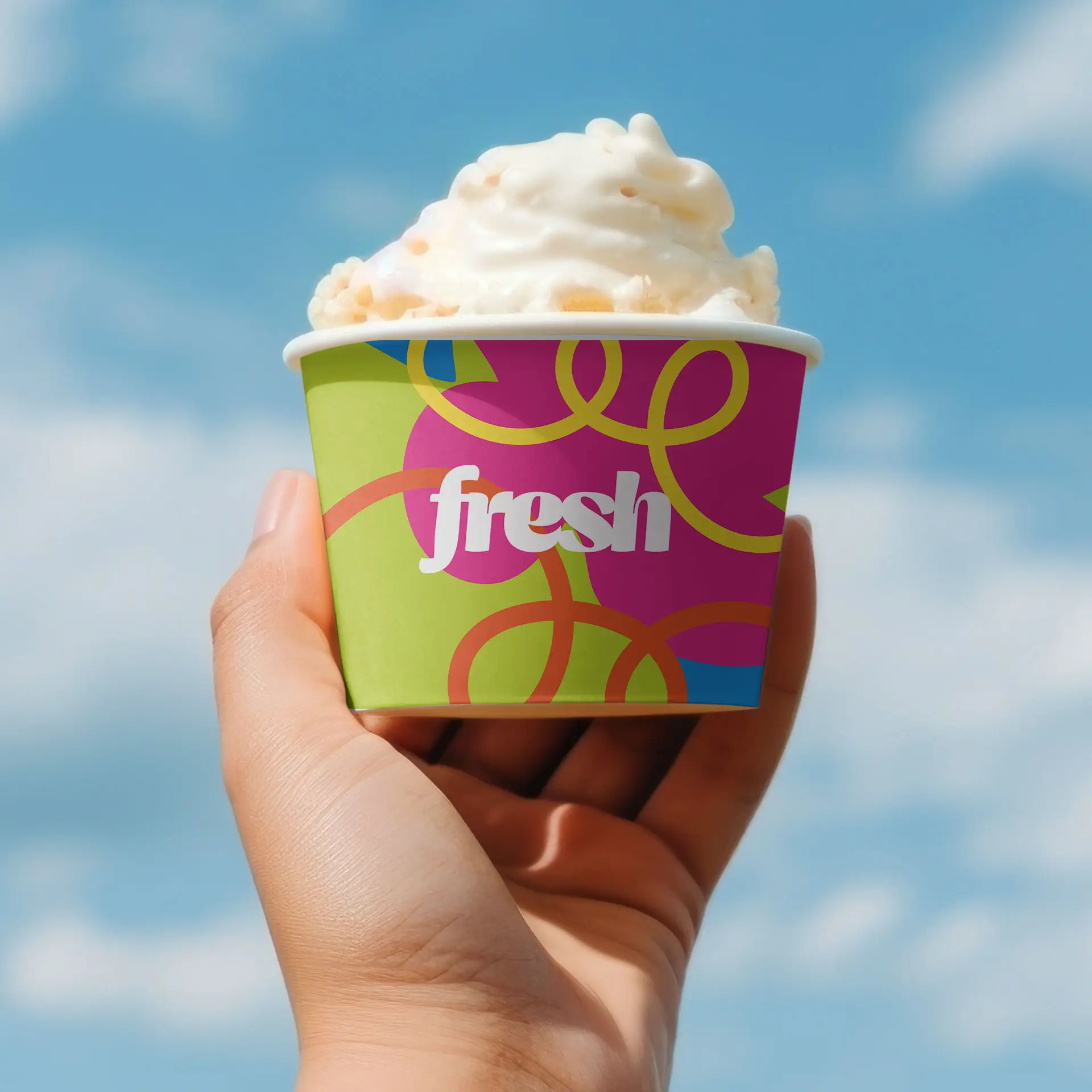 Hand holding a colorful ice cream cup with a lid, featuring the word "fresh" and topped with creamy ice cream against a blue sky.