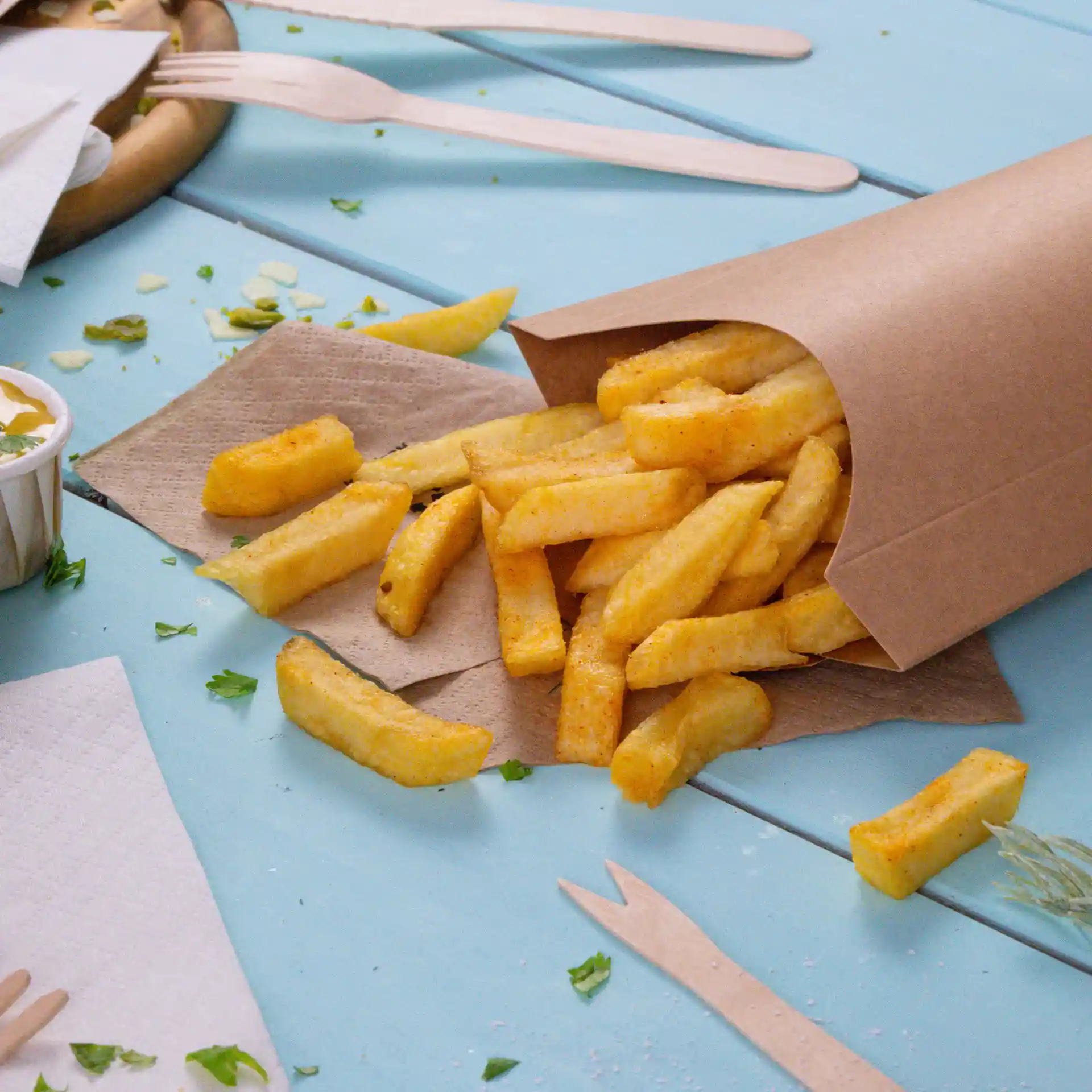 Brauner Papierbecher gefüllt mit goldenen Pommes, auf einer hellblauen Oberfläche mit verstreuten Servietten und Holzbesteck.