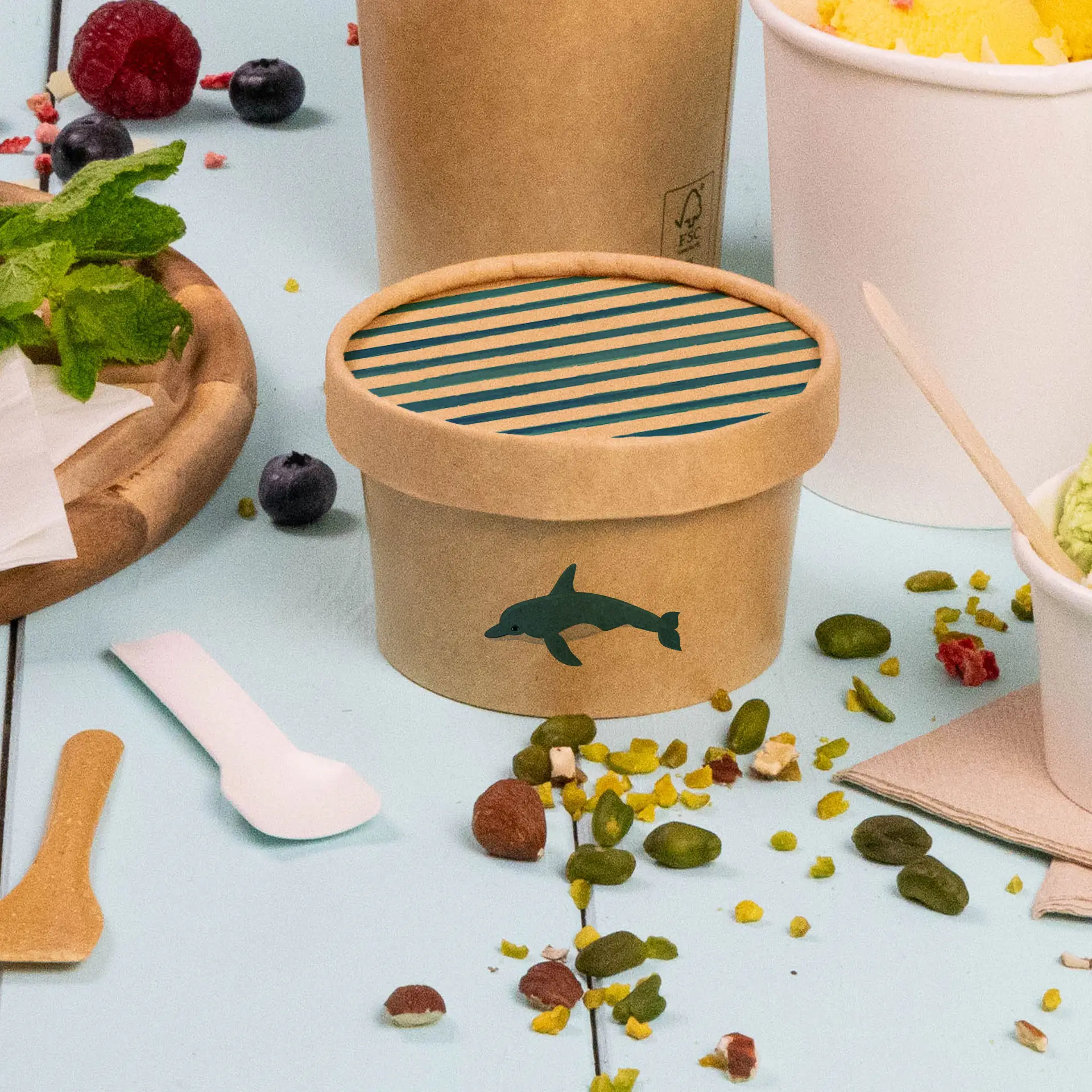 One brown cardboard lid for ice cream cup with a dolphin design and striped blue interior; surrounded by toppings and utensils.