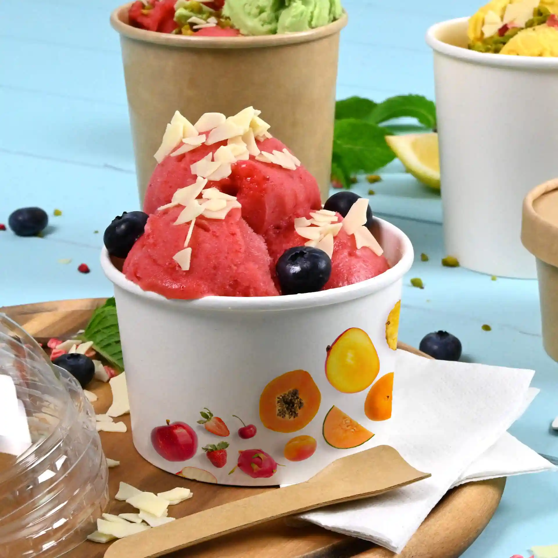 Round white ice cream cup with pink sorbet, topped with almond slivers and blueberries, on a wooden tray with fruit images.