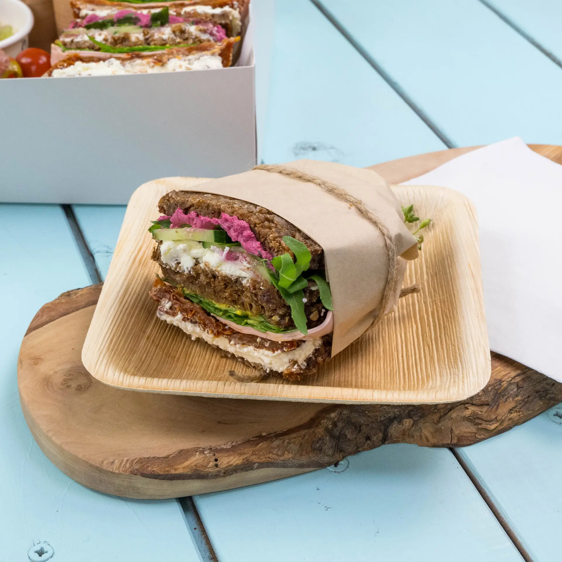Braun eingewickeltes Sandwich auf einem Holzbrett, mit Schichten von Grünzeug, Frischkäse und rosa Aufstrich.