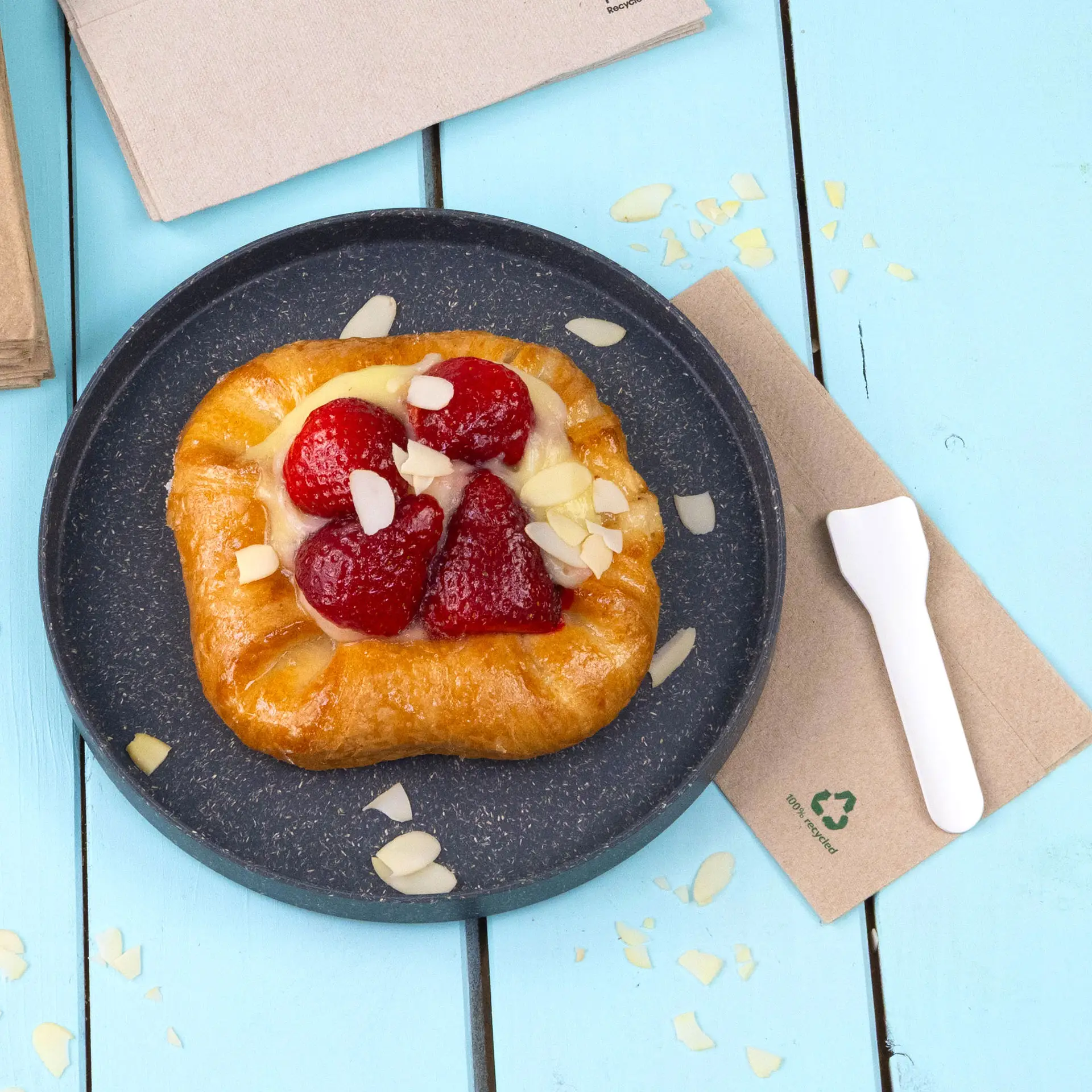 Pastry topped with strawberries and almond flakes on a black plate, surrounded by paper napkins on a blue background.