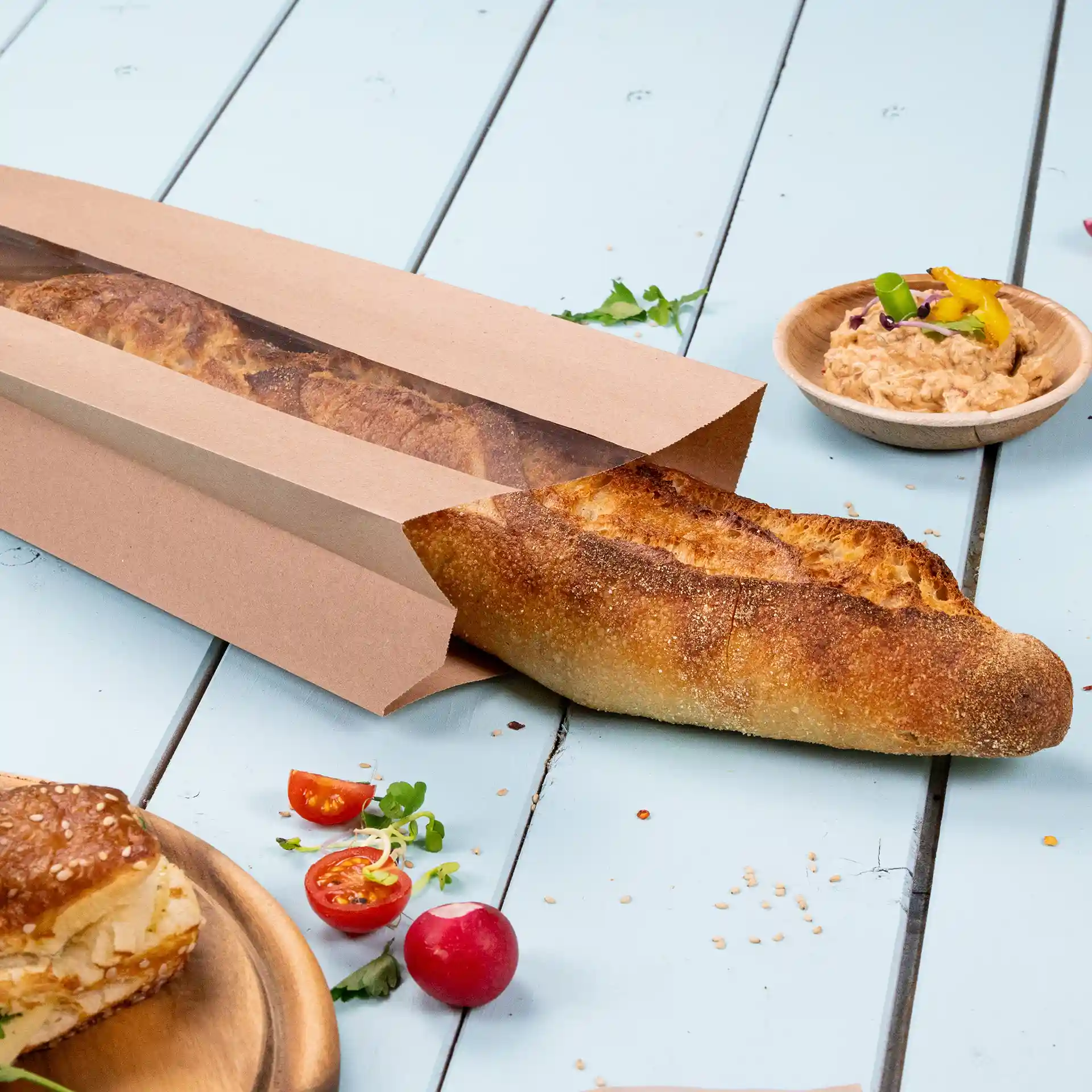 Braune flache Papiertüte mit Fenster, die ein Baguette enthält, auf einer hellblauen Oberfläche mit Tomaten und Dip.