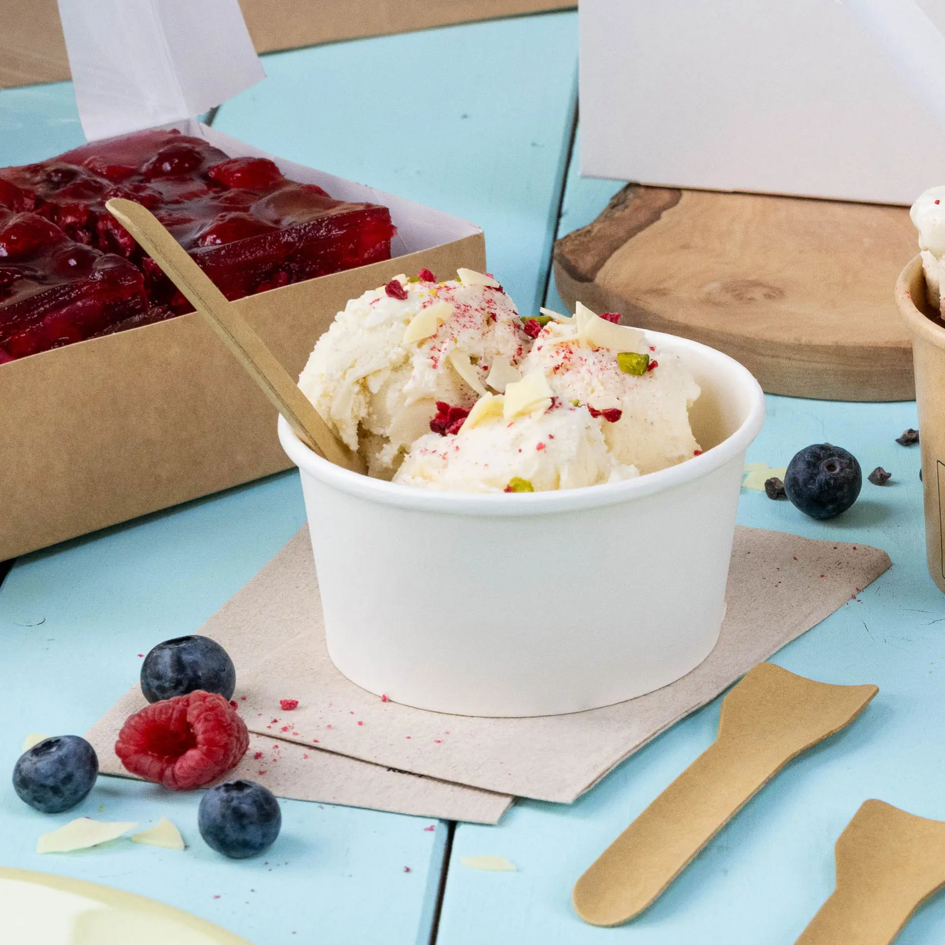 Becher mit Eis, garniert mit Obst und Nüssen, Holzlöffel und frischen Beeren auf blauer Oberfläche mit Serviette.