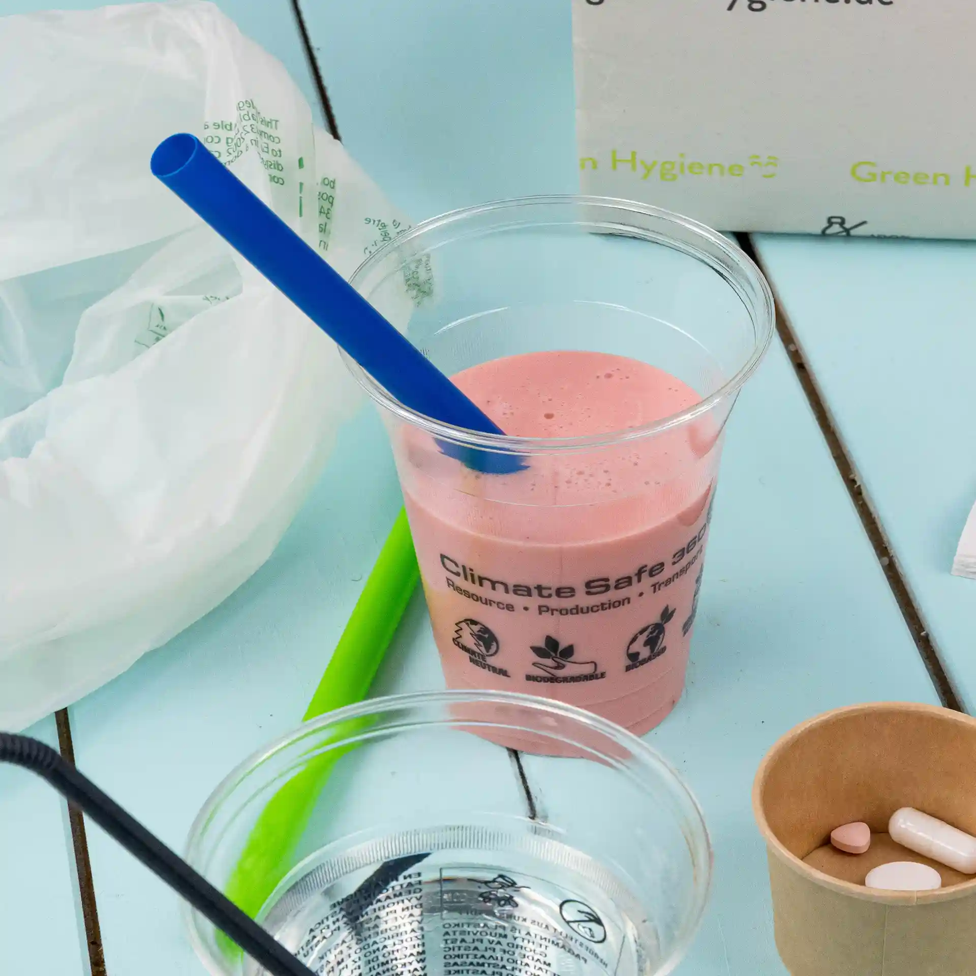 Image of a clear cup with pink beverage and a blue straw, alongside a green straw, a glass of water, and a paper cup with pills.
